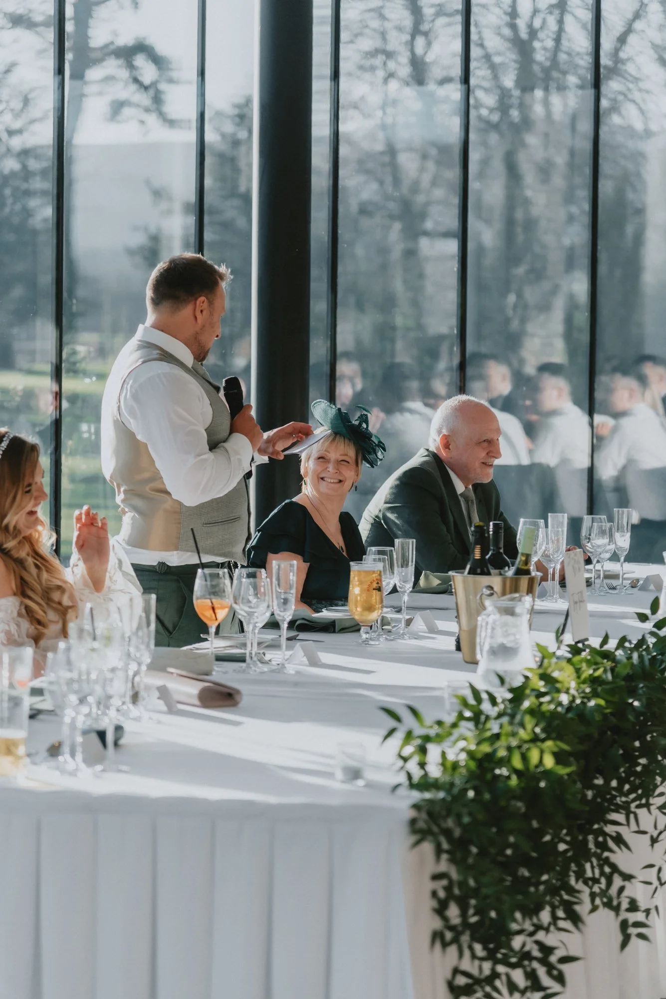 A man giving a toast at a wedding banquet, with happy guests seated at a long decorated table inside a glass-walled venue, with trees outside.