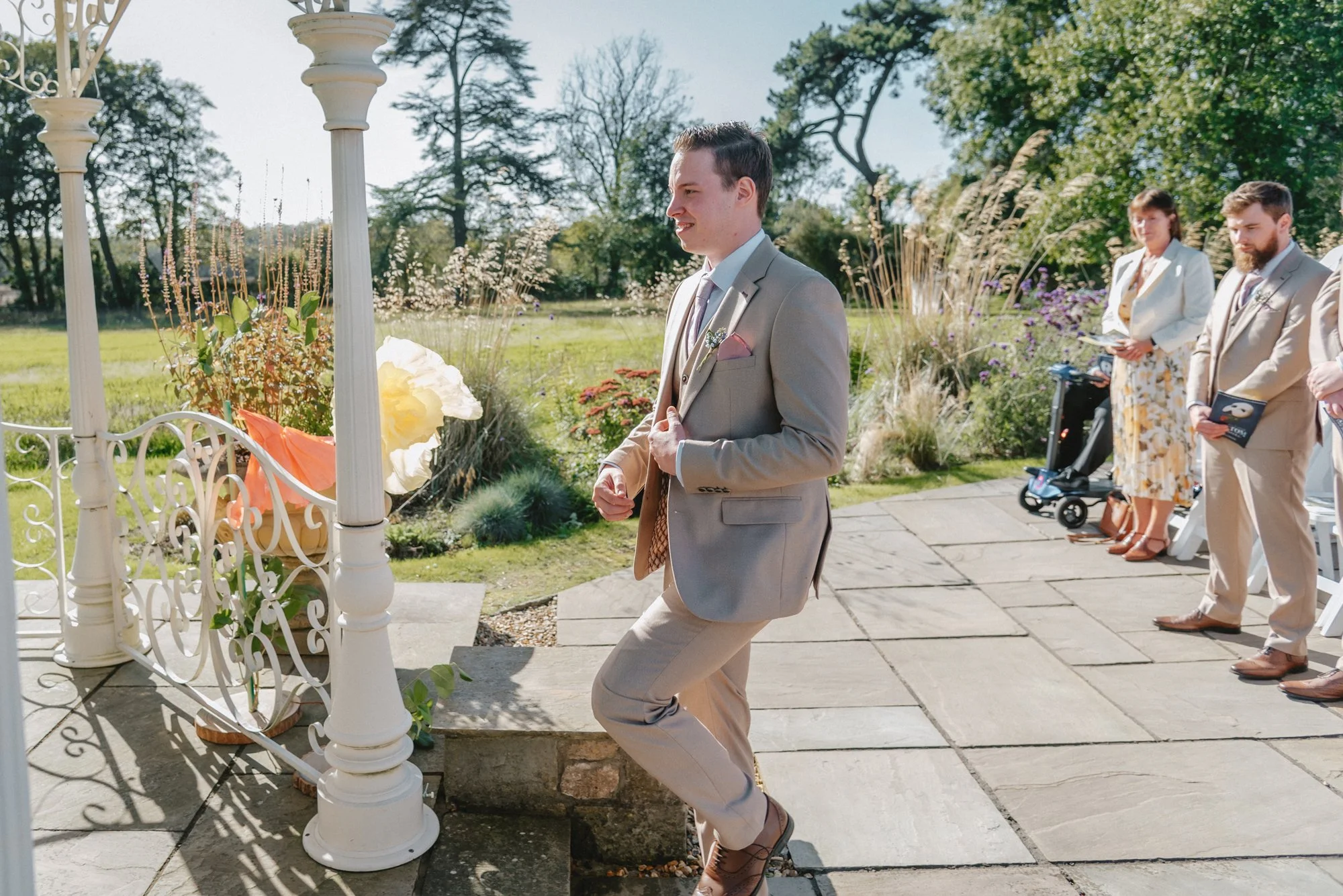 A man in a light gray suit dancing at an outdoor wedding ceremony with guests standing nearby, greenery, and floral decorations in the background.