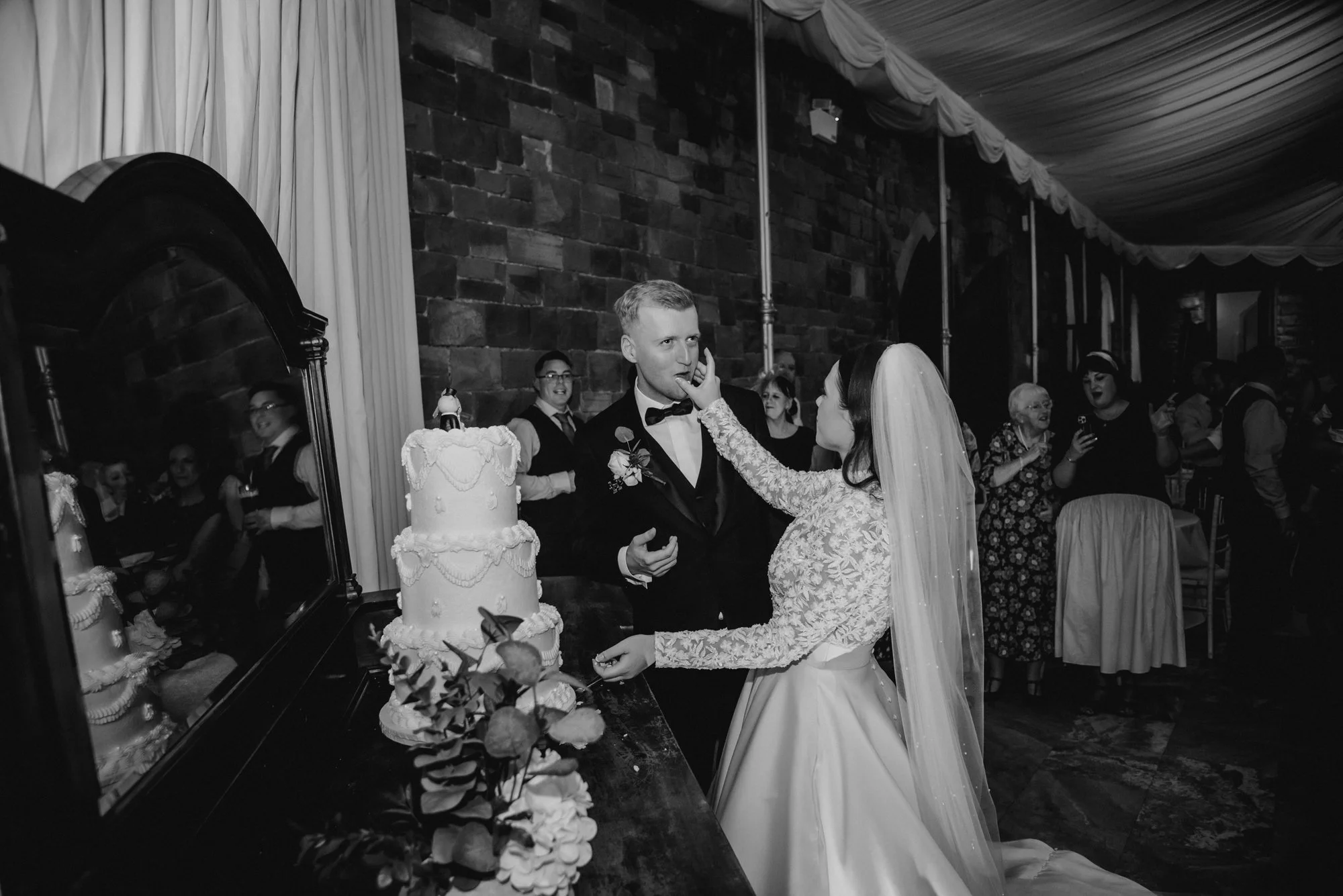 Bride and groom at their wedding cake table, with the bride feeding the groom a piece of cake, surrounded by wedding guests, in a decorated indoor venue.