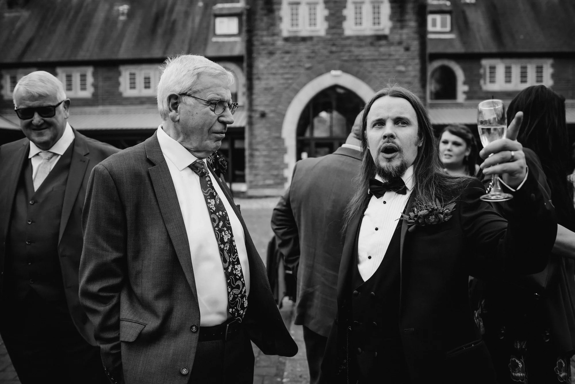 A black and white photo of a formal gathering outside a building with a brick and arched entrance. Several people are dressed in suits and tuxedos, including a man with long hair and a beard raising a glass in a toast and an older man wearing glasses