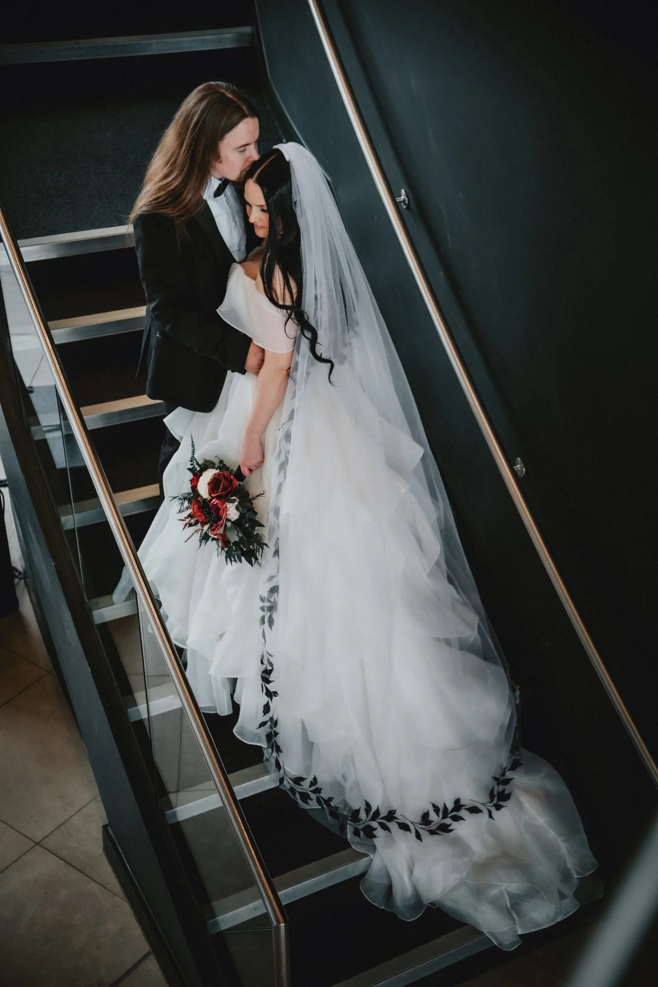 A bride and groom share a kiss on a staircase. The bride wears a white wedding gown with black leaf embroidery along the hem and a long veil. The groom wears a black suit with a white shirt. The bride holds a bouquet of red and white flowers.