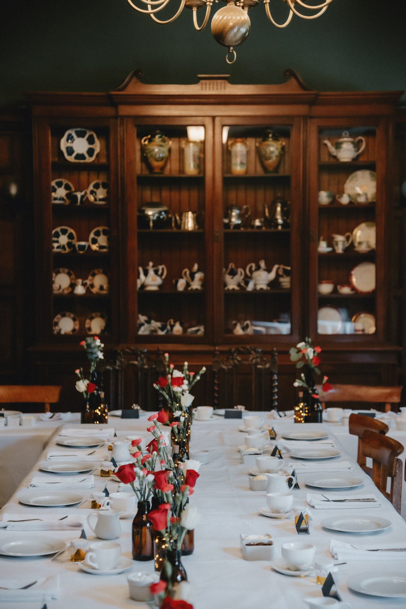 A dining table set for a formal event with white tablecloths, plates, cups, and cutlery. Small vases with red and white flowers decorate the center. A large wooden china cabinet filled with various china, teapots, and silverware is in the background 
