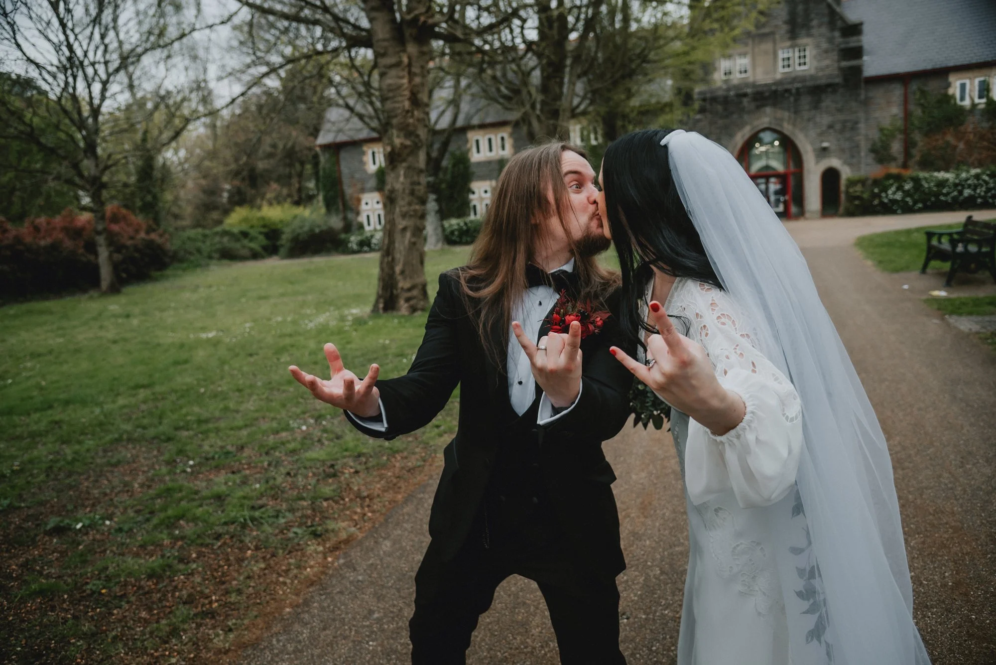 A couple dressed as a groom and bride sharing a kiss outdoors on a wedding day, with the groom wearing a black tuxedo and the bride in a white lace dress and veil.