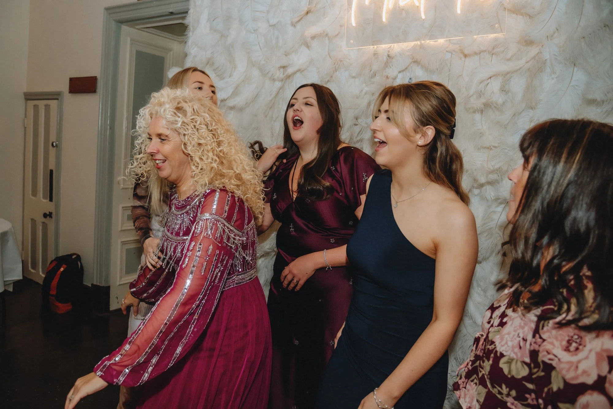 A group of women enjoying a conversation at a social event with a feathered white wall background.