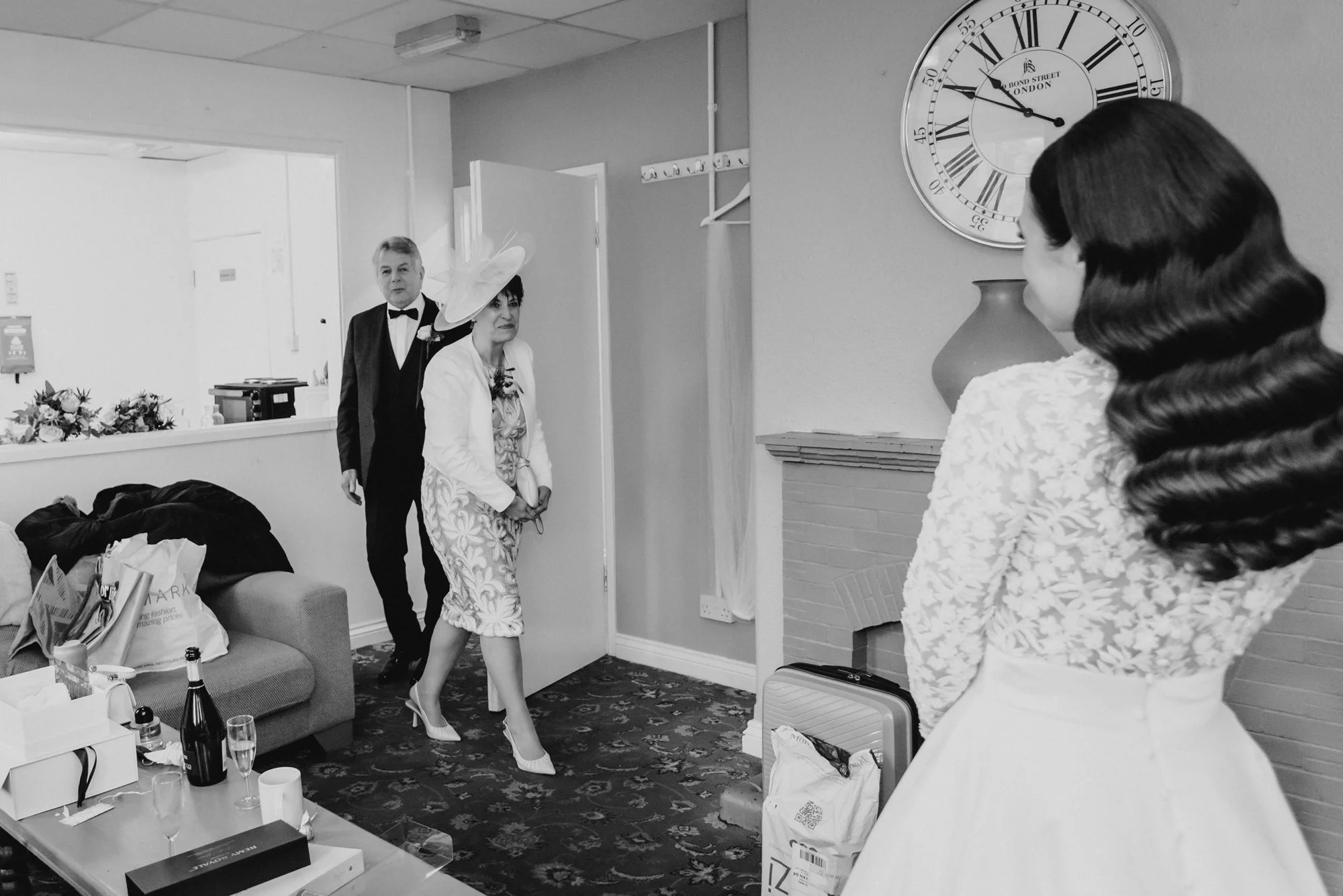 A black and white photo of a woman with long, wavy hair, dressed in vintage clothing, looking at two people entering through a door. The woman entering is wearing a floral dress, white blazer, heels, and a large hat. The man entering wears a tuxedo w
