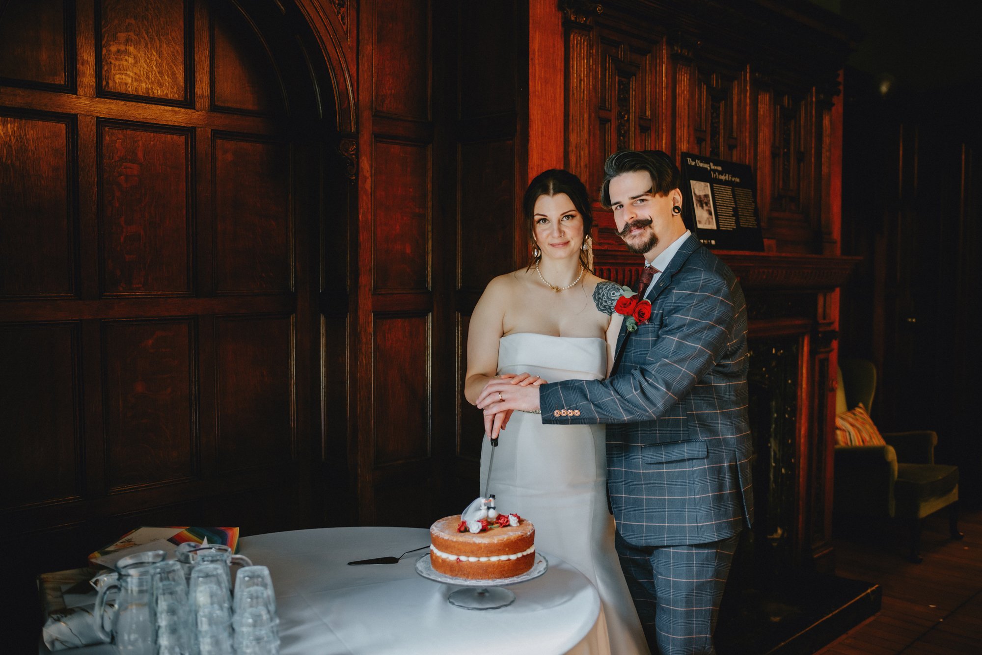 A couple at a wedding or celebration, standing behind a table with a cake. The woman wears a white strapless dress and the man wears a gray plaid suit with a red rose boutonniere. They are holding hands and smiling at the camera in a wood-paneled roo