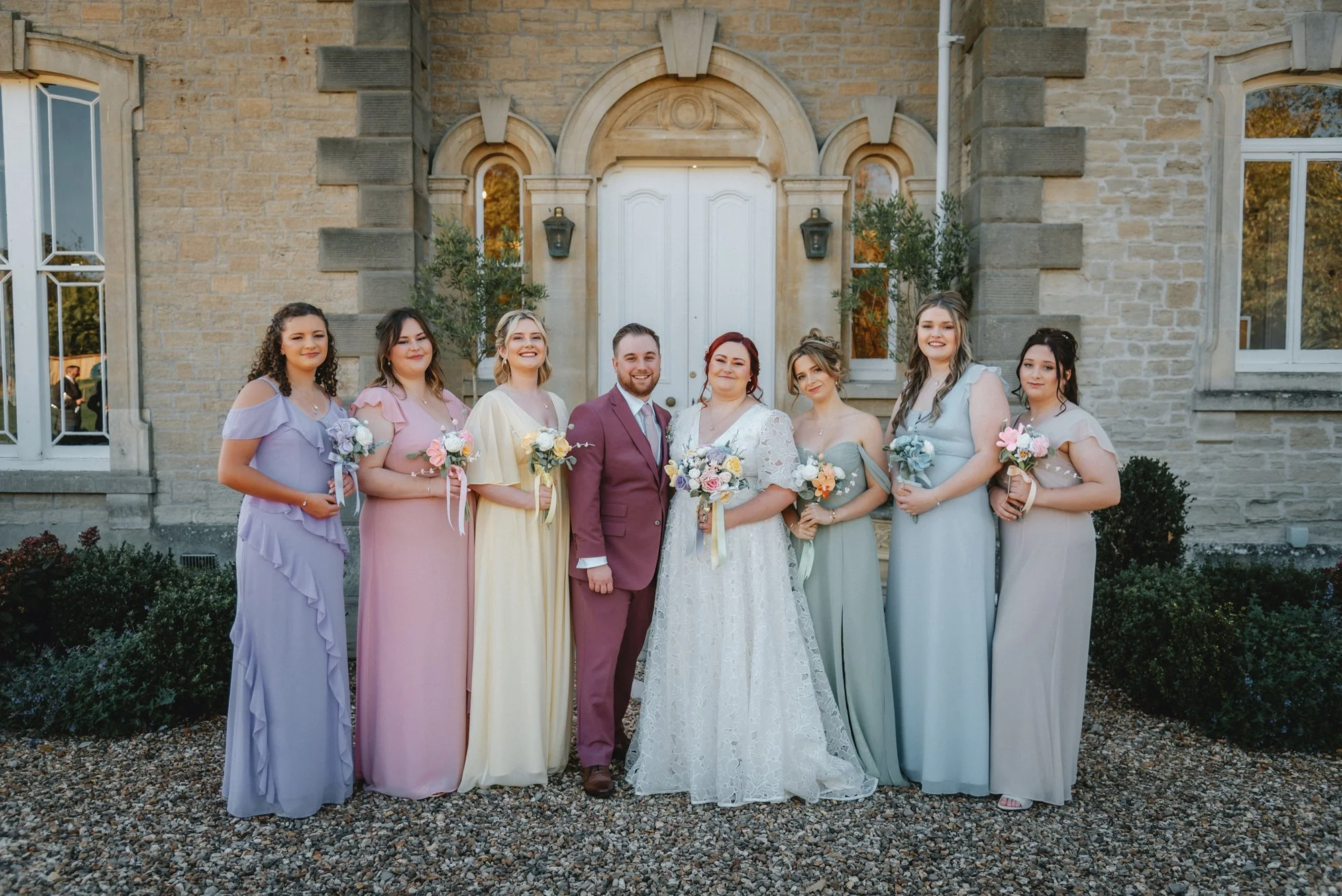 A wedding party standing outside a stone building with arched windows and a white door. The group includes the bride and groom with six bridesmaids, all holding bouquets, dressed in pastel and neutral colors.