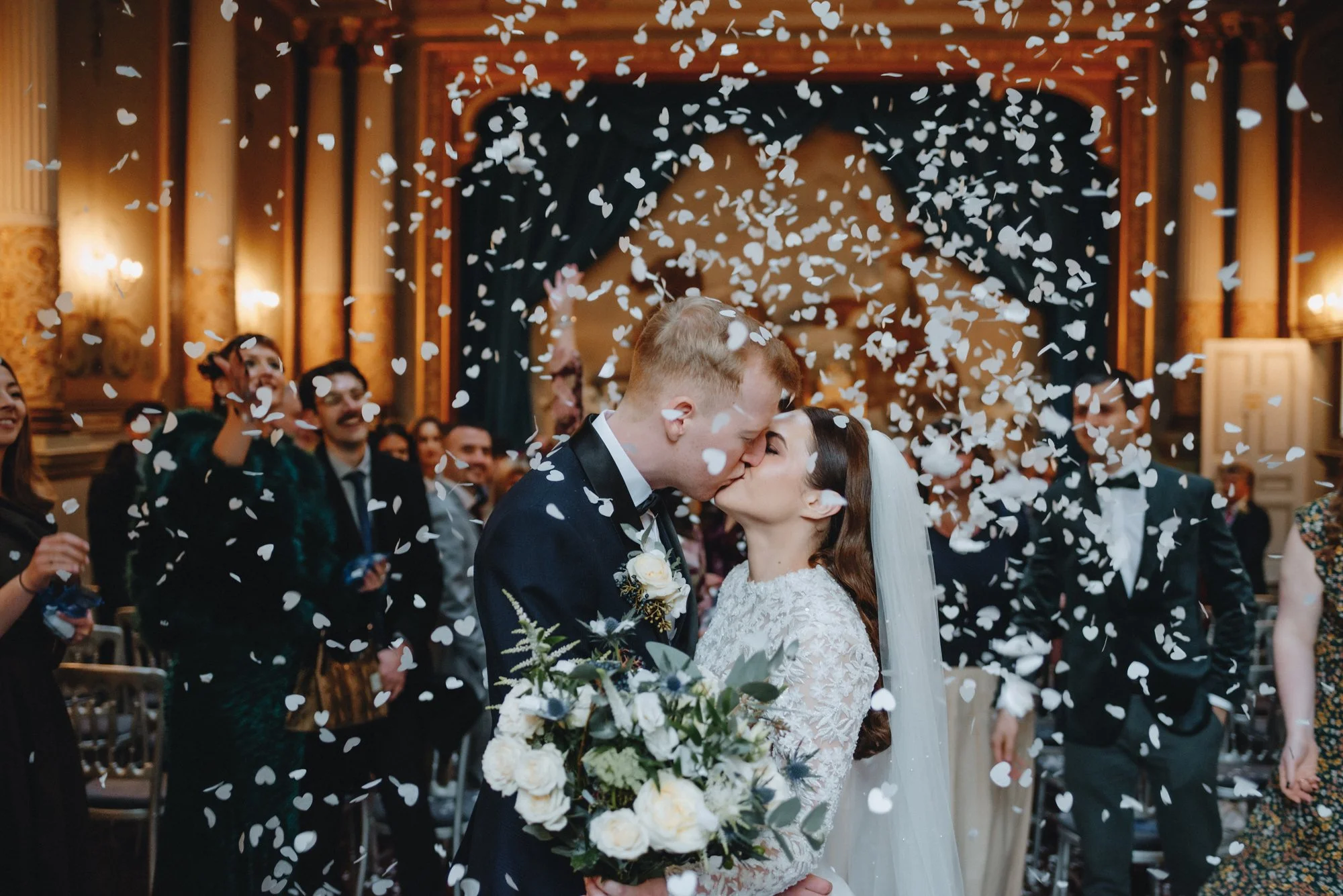 A newlywed couple kisses under falling white confetti in a decorated wedding hall, surrounded by friends and family.