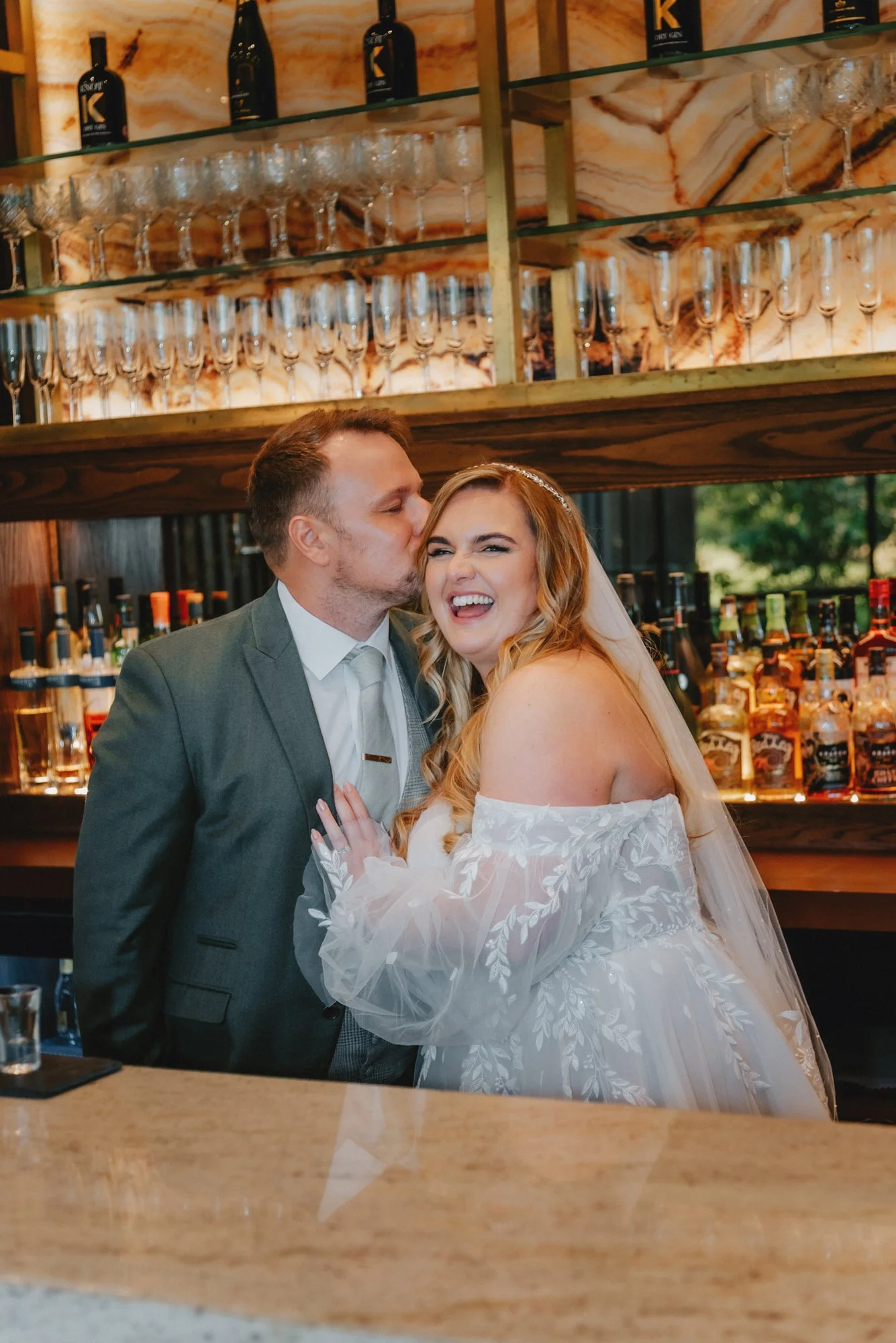 A newlywed couple standing at a bar, with the groom kissing the bride on the cheek. The bride is laughing joyfully, wearing a white off-the-shoulder wedding dress with floral details. The bar has shelves with glasses and bottles behind them.