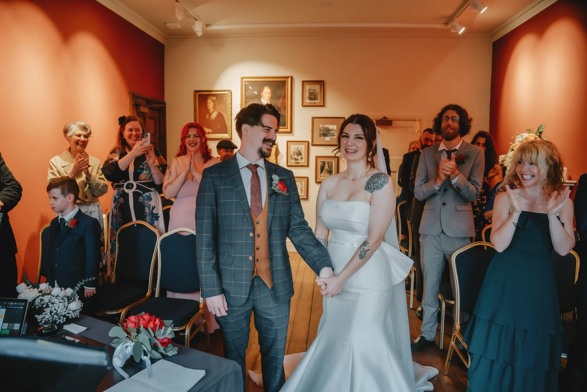 A bride and groom holding hands during their wedding ceremony surrounded by smiling guests in an art gallery with framed portraits on the wall.