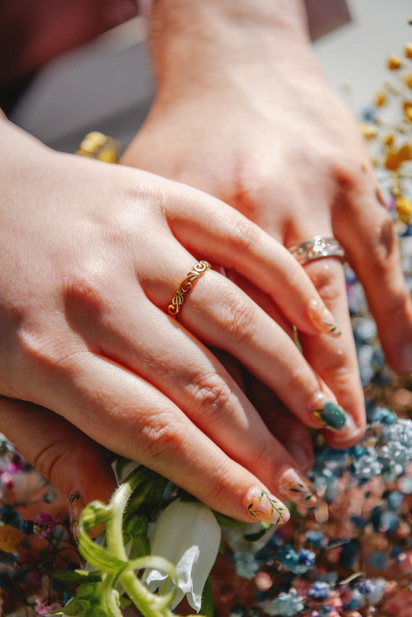 Close-up of a person's hands with rings, one with a gold ring with a swirl pattern and another with a silver ring, resting on colorful flowers. The person has manicured nails with floral designs.
