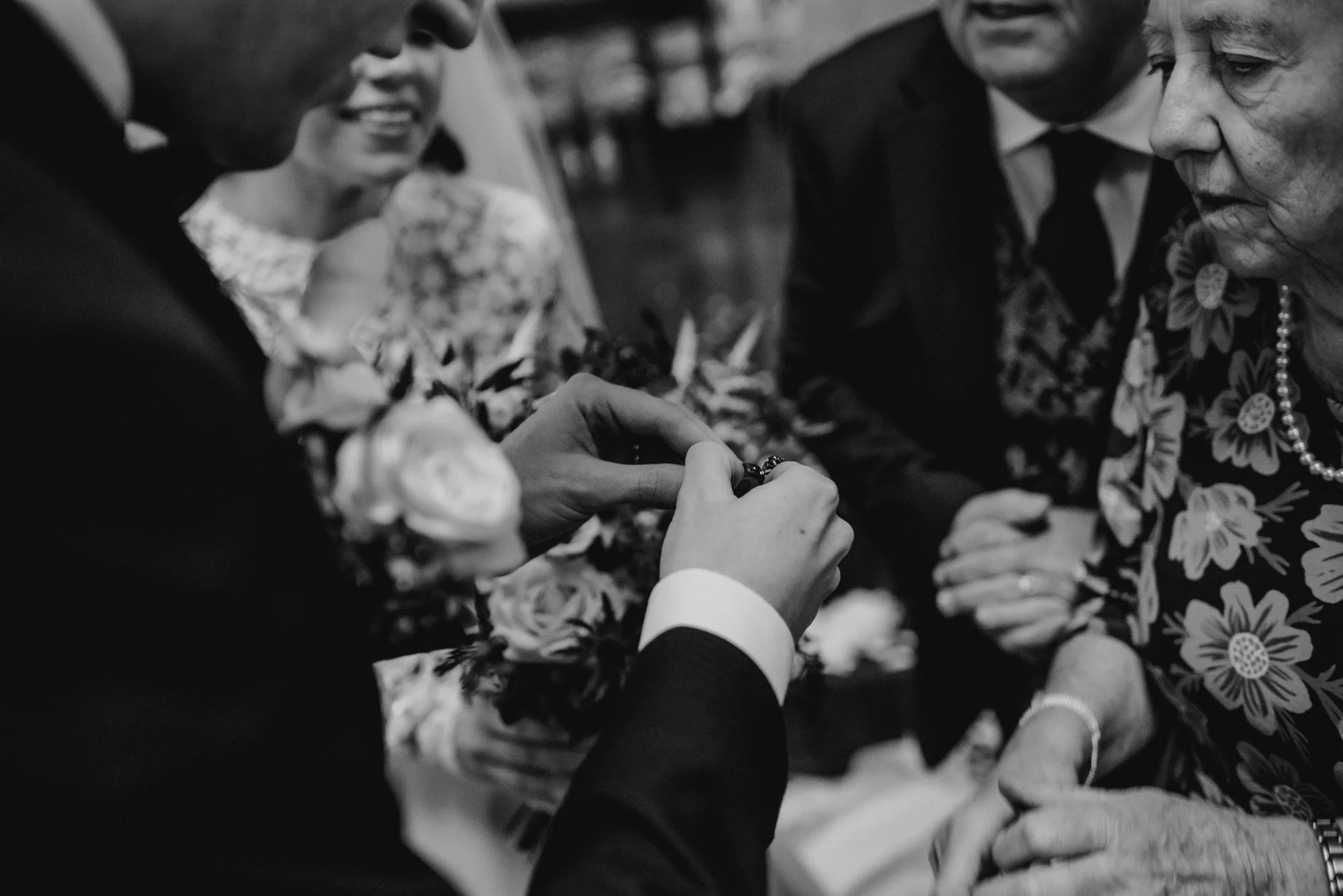 A close-up of a wedding or ceremonial ceremony where a person is helping another person with a ring or jewelry, surrounded by three other women and a man, all dressed formally, in a black and white photo.