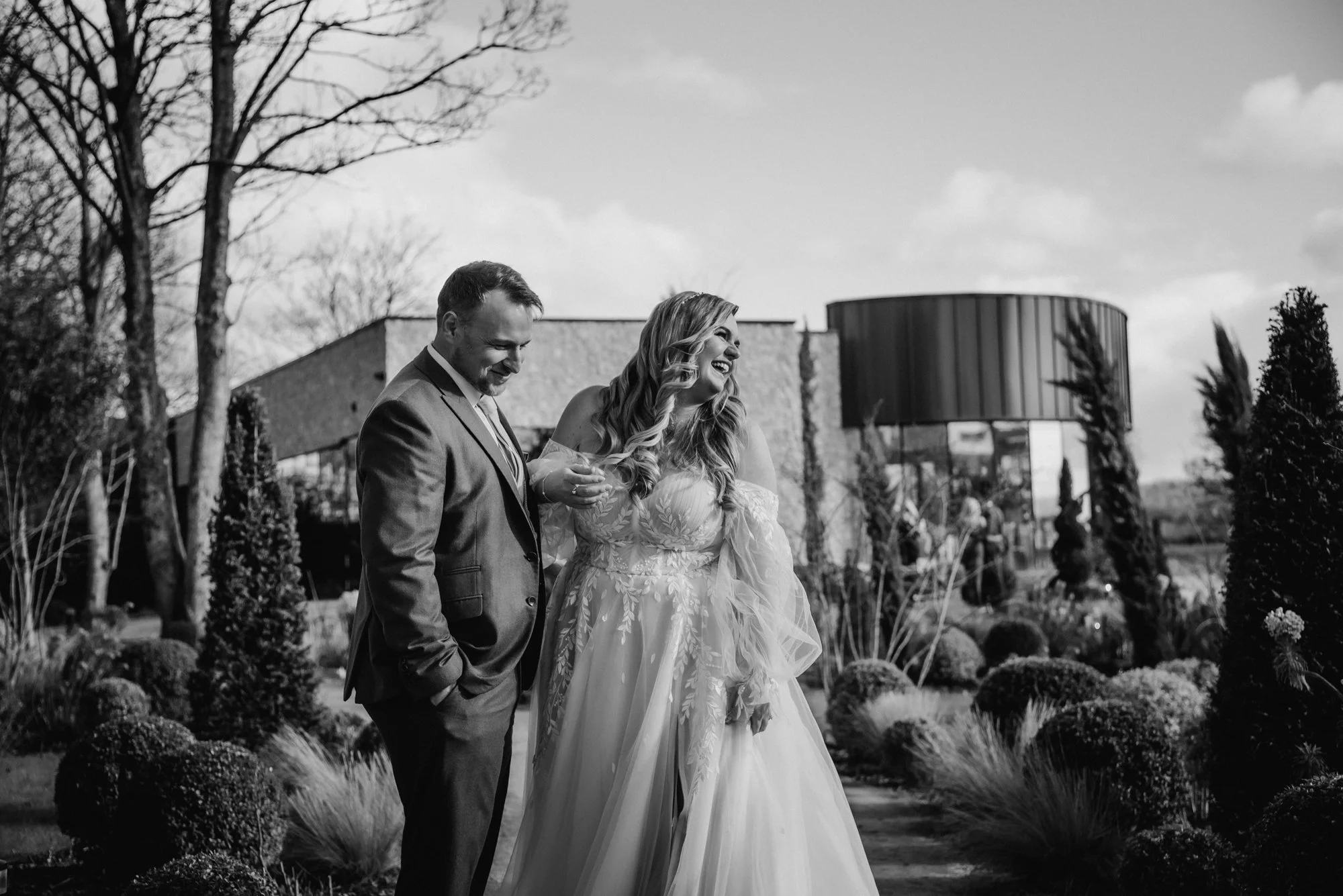 A black and white photo of a bride and groom standing outdoors, smiling and sharing a moment together, with a modern building and trees in the background.