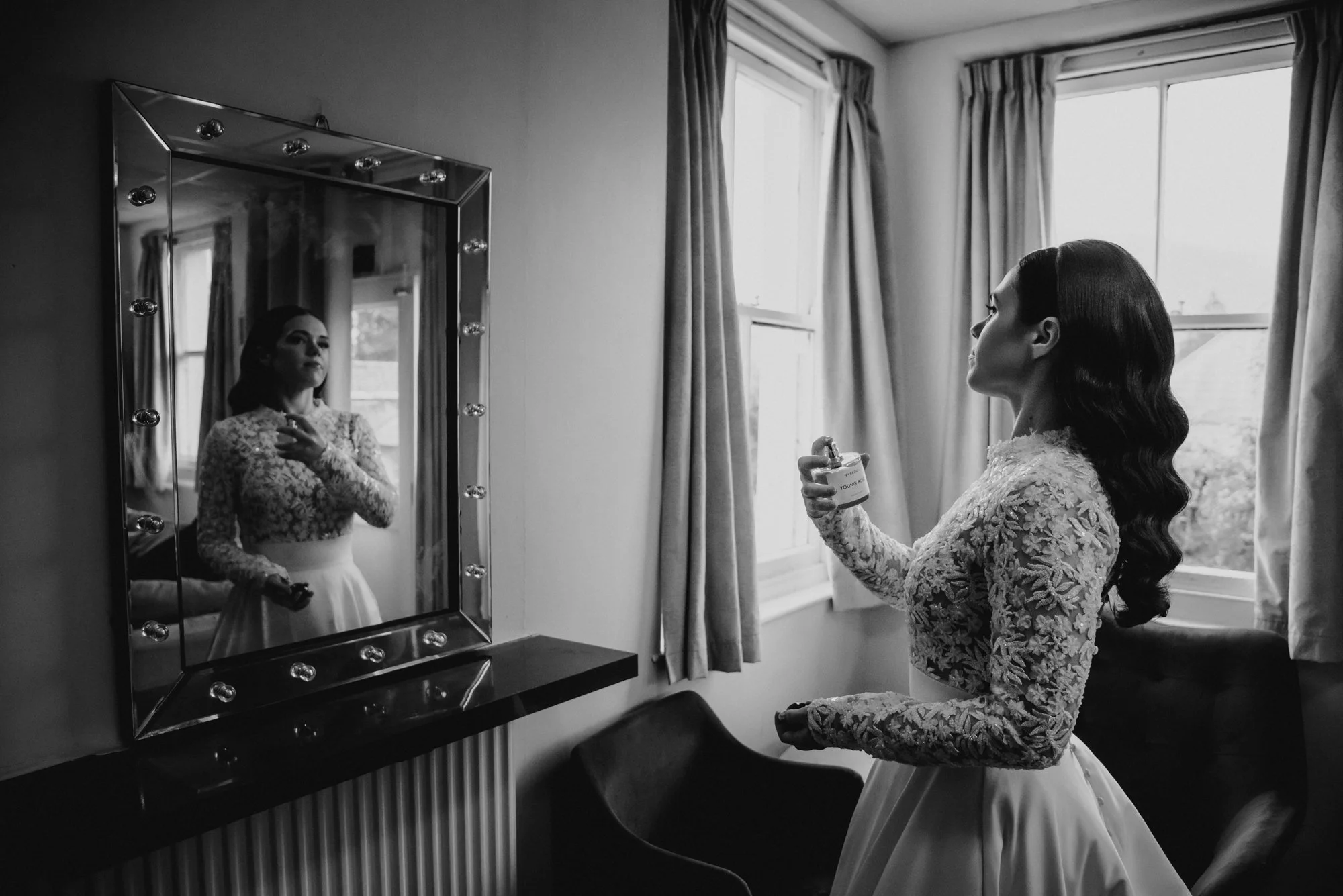 A woman in a lace dress looks in a mirror while holding a perfume bottle, with sunlight coming through the window behind her.