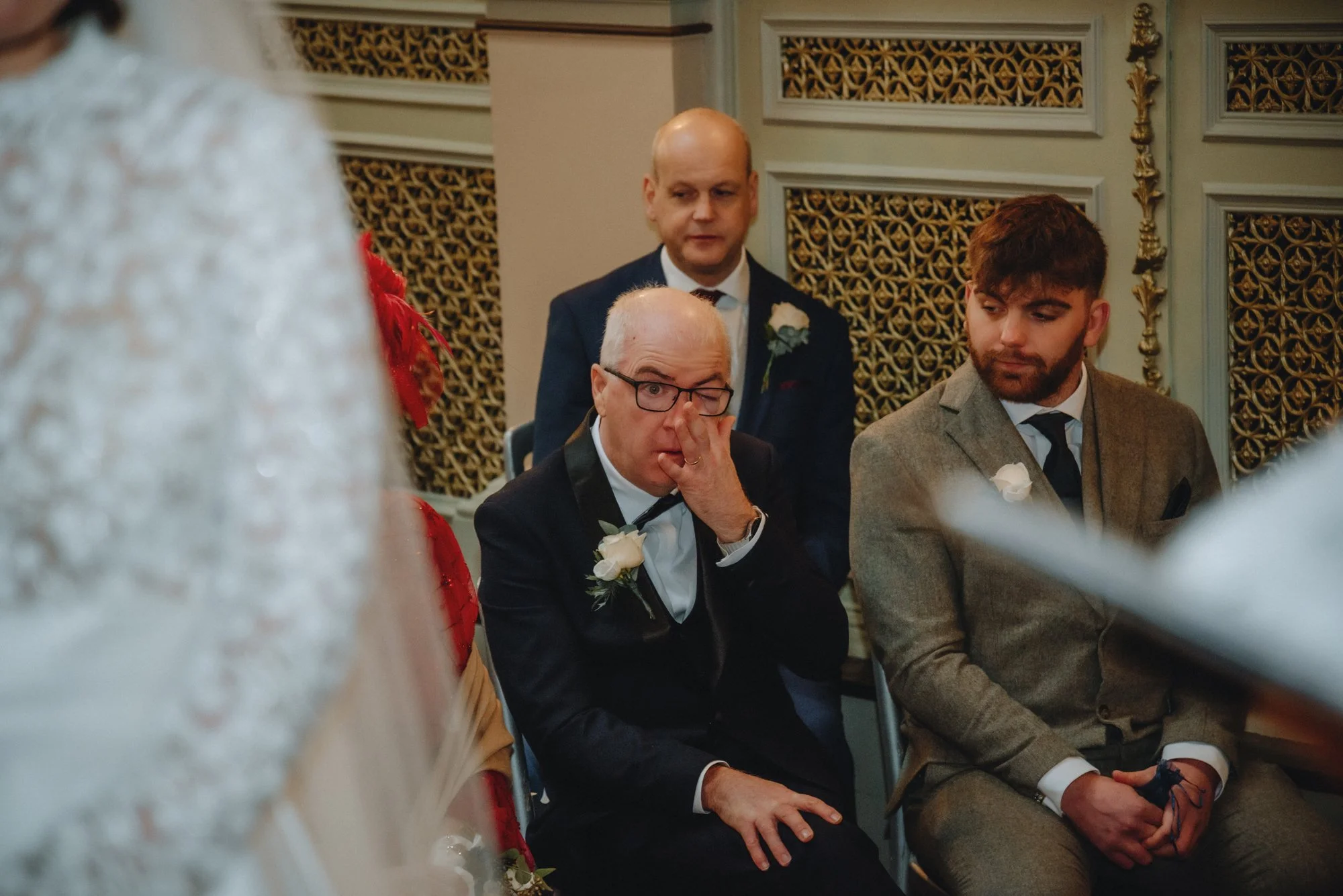 Three men in formal attire sitting at a table during a wedding event. The man in the foreground is old with glasses, wearing a black suit and white boutonniere, covering his face with his hand. The man on the right has a beard, wearing a gray suit an