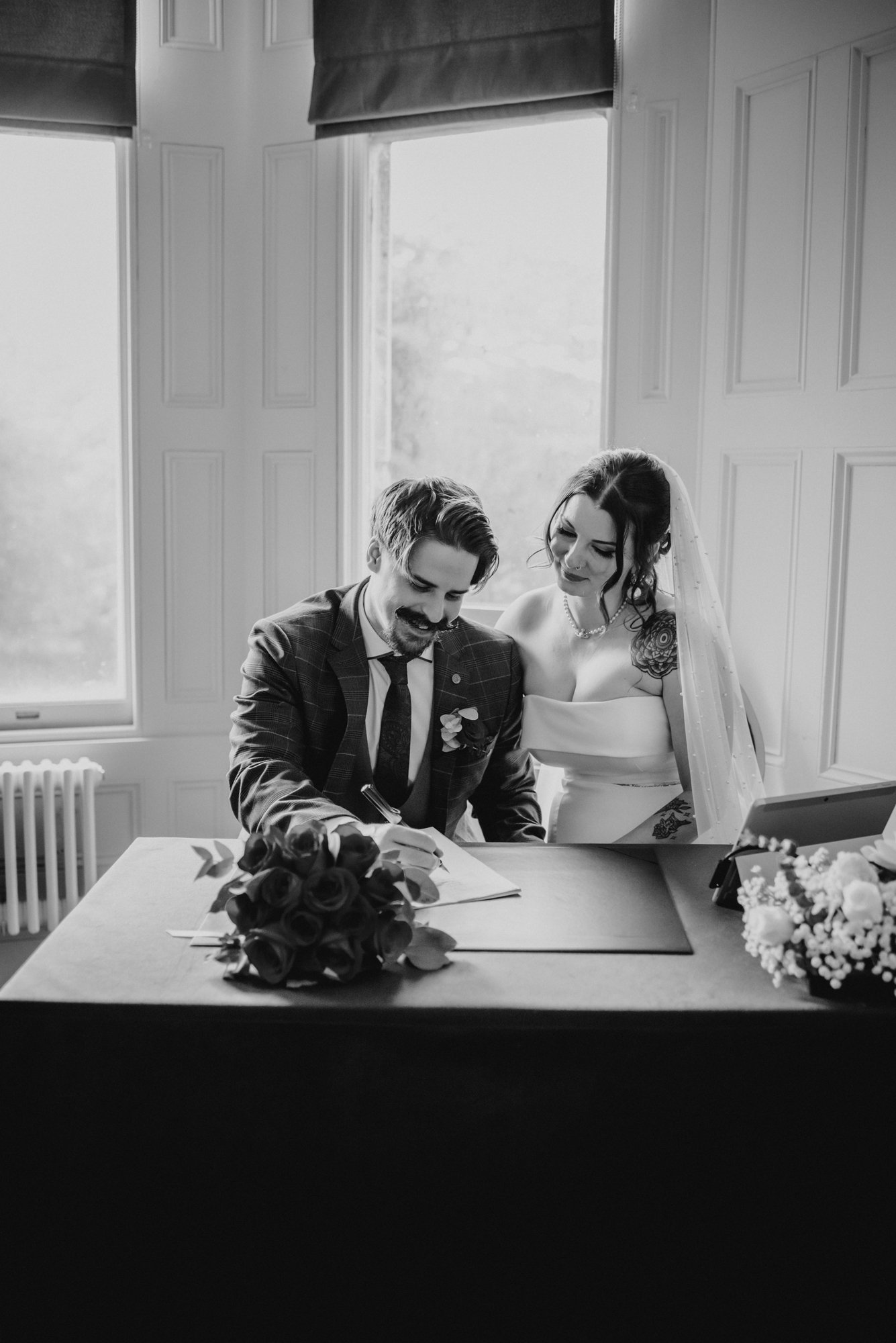 A black-and-white photo of a bride and groom signing a document, with the bride smiling and the groom focused on the paper, a bouquet of flowers on the table.