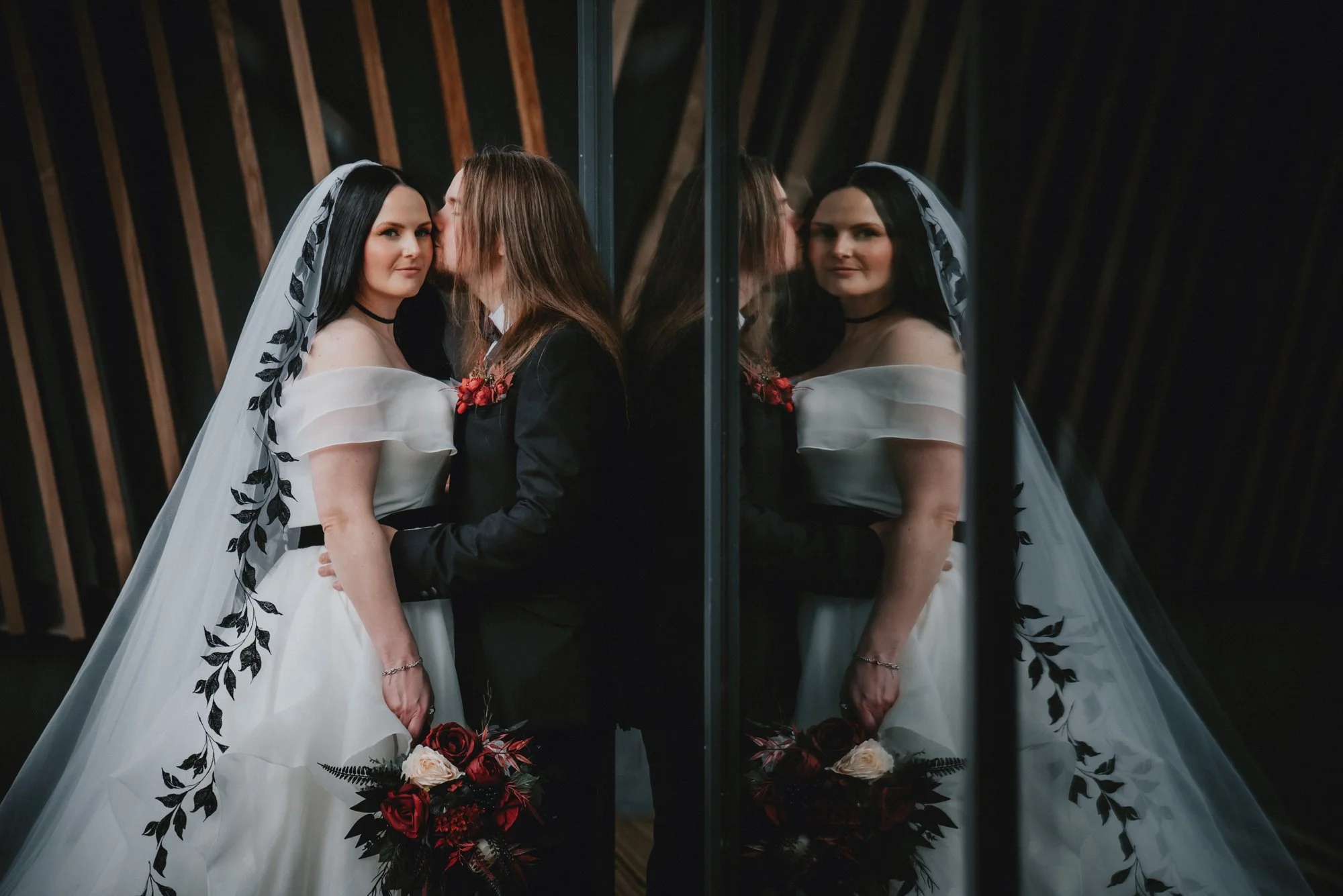 A bride and groom stand close together in front of a mirror, with the bride wearing a white wedding gown with black floral details and a veil, and the groom in a dark suit with a red floral boutonniere. The bride holds a bouquet of red and white rose