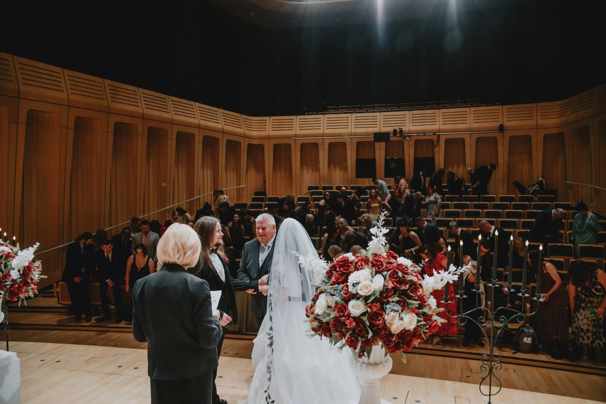 A wedding ceremony taking place in a concert hall or theater with wooden walls. The bride, wearing a white wedding gown and veil, is holding hands with the groom, who is dressed in a suit. They are standing at an altar adorned with large floral arran