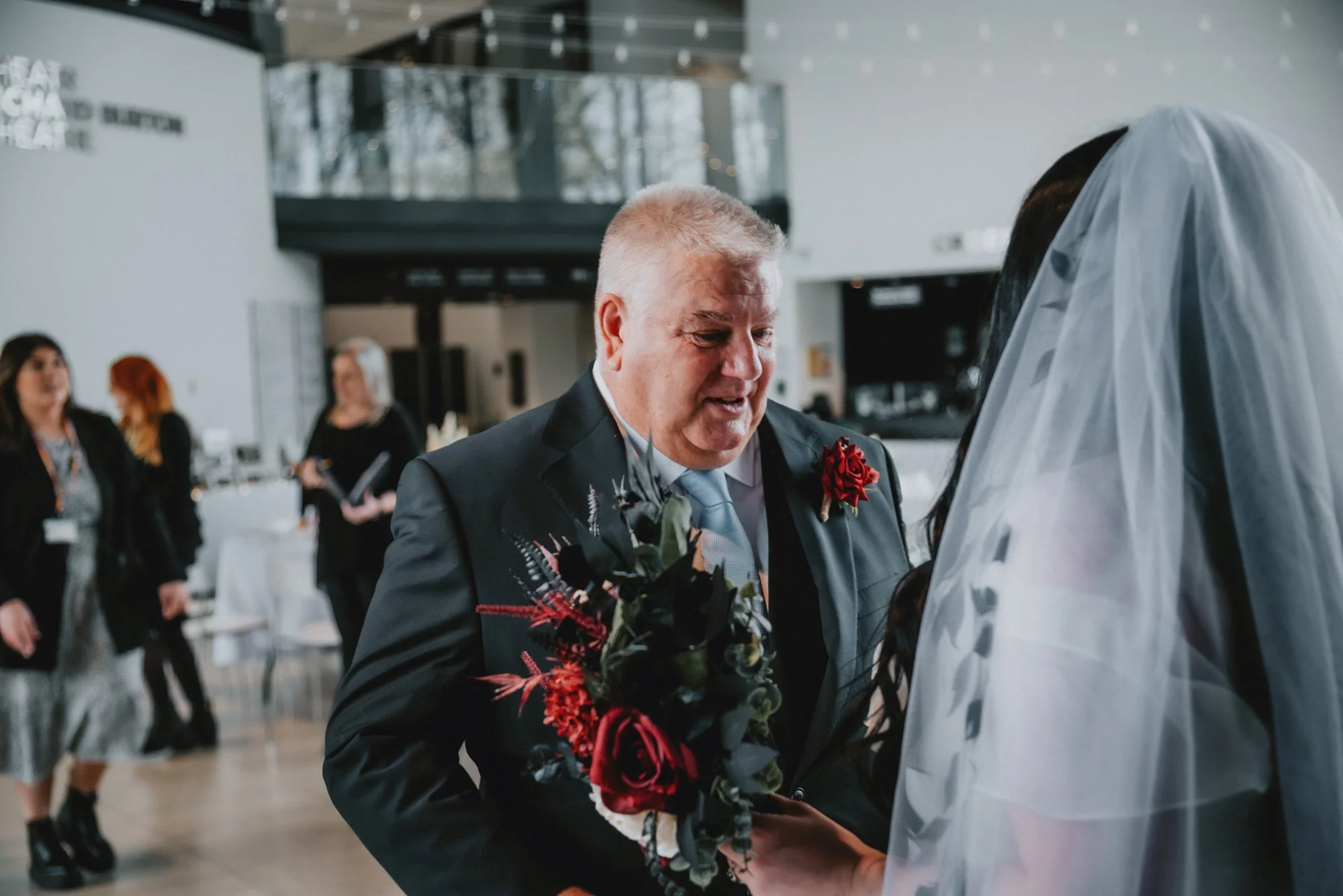 A heartfelt moment during a wedding ceremony where an older man in a suit with a red flower boutonniere is holding the hands of a bride dressed in white with a veil, in a modern indoor venue.