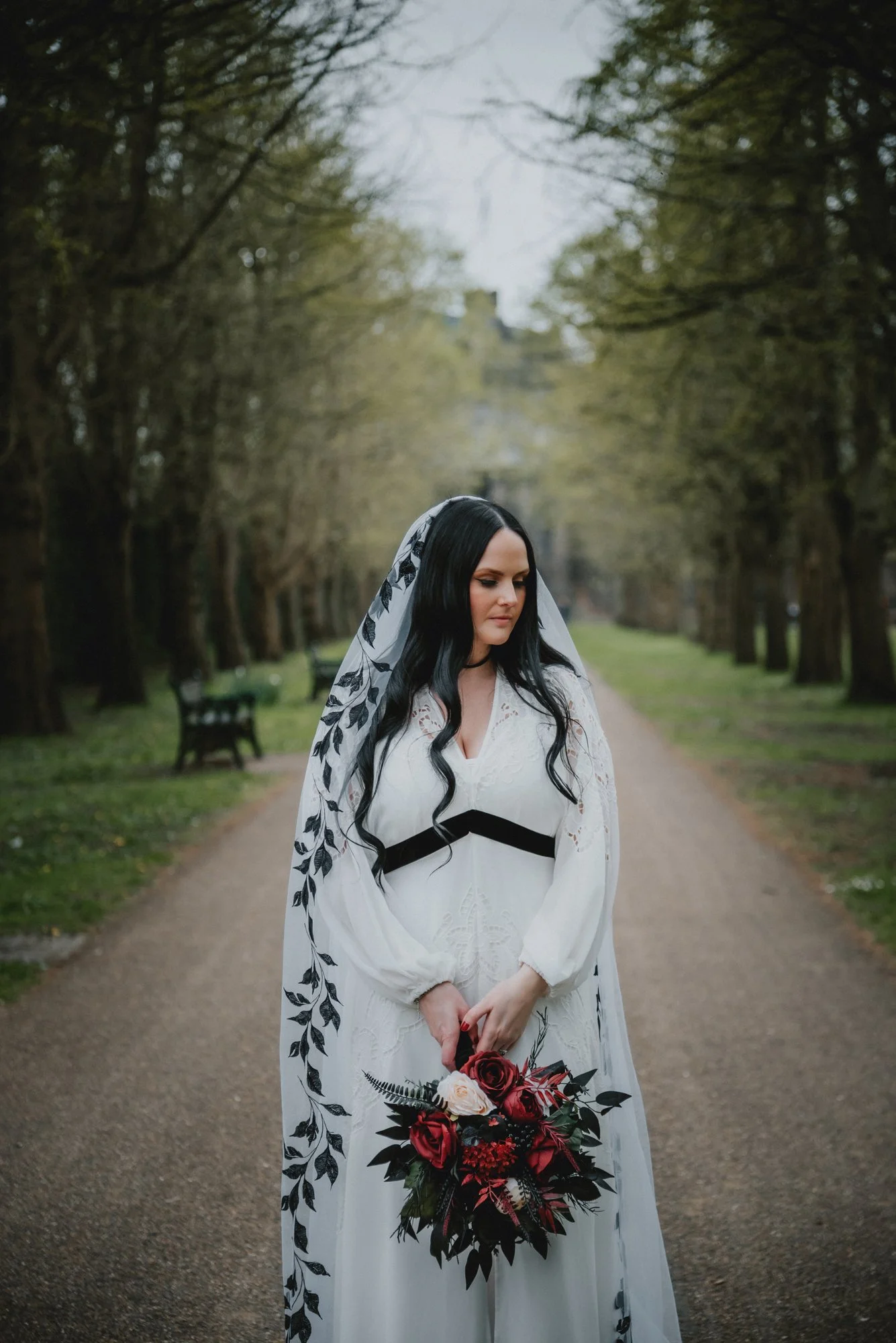 A woman dressed in a white wedding gown with black accents, holding a bouquet of red and white roses and greenery, standing on a pathway lined with trees in a park.
