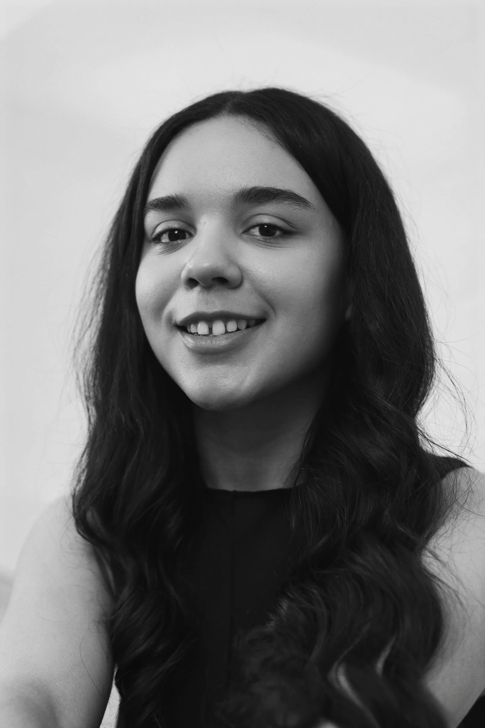 Black and white portrait of a young woman with long wavy hair, smiling, wearing a sleeveless top, against a plain background.
