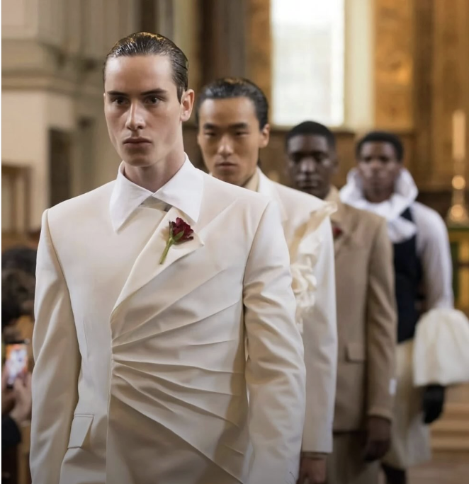 Four men standing in a line, dressed in formal cream-colored suits, inside a church or historic building, with one man in front wearing a boutonniere, looking serious.