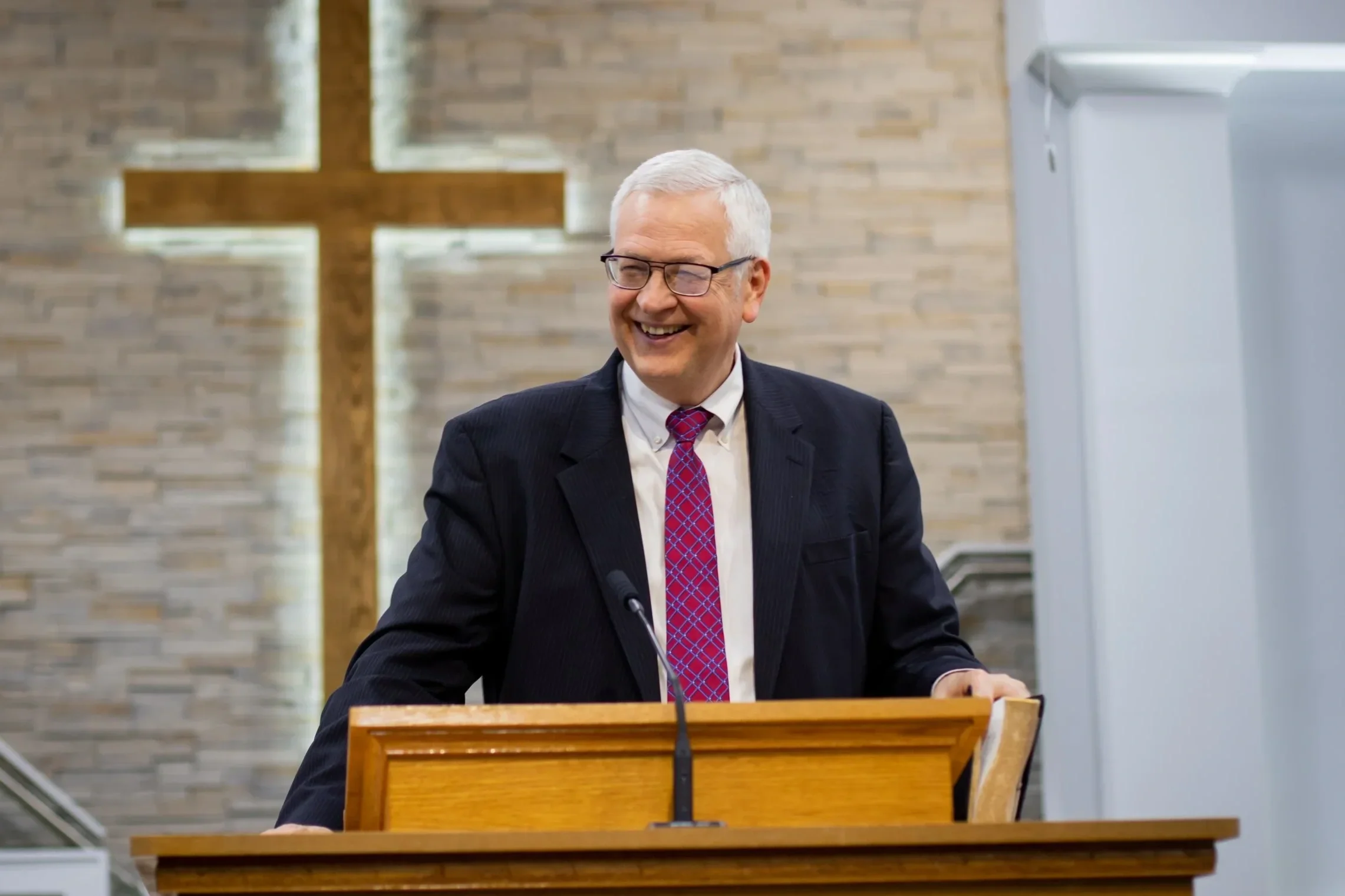 A smiling man in a black suit, white shirt, and red patterned tie, standing behind a wooden pulpit with a microphone, inside a church with a lit cross on the brick wall behind him.
