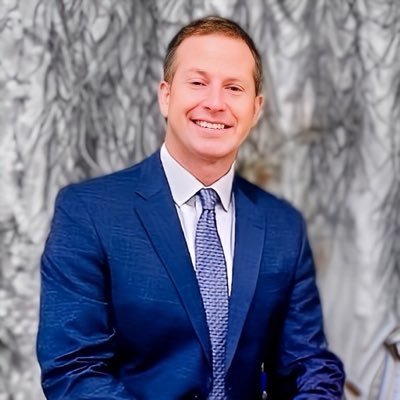 A man in a blue suit and tie smiling in front of a gray, textured background.