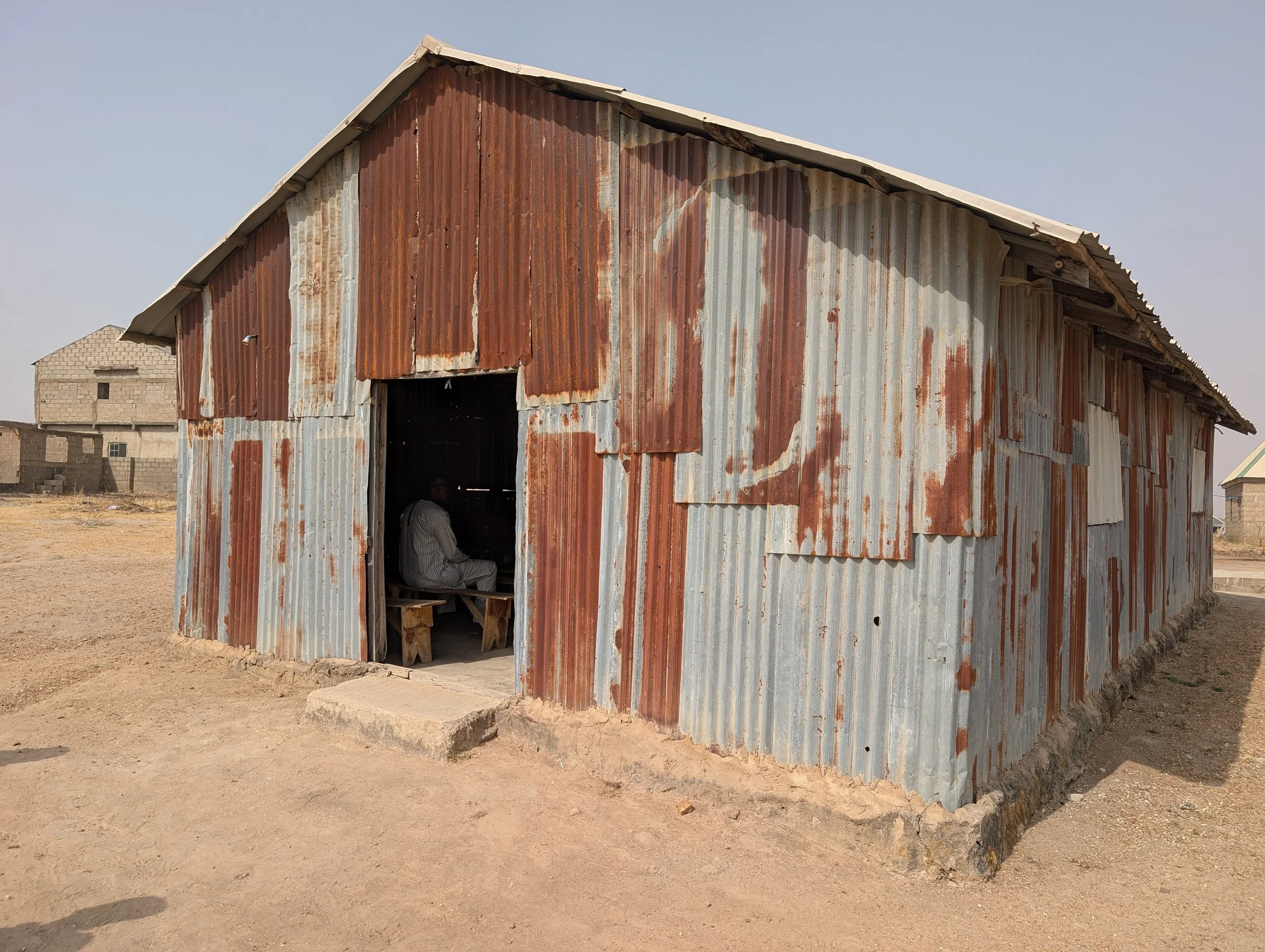 Rusty metal shed with open door, a person sitting inside, and a dry, dirt ground outside.