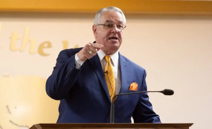 A man in a blue suit and yellow tie speaking at a podium with a microphone.
