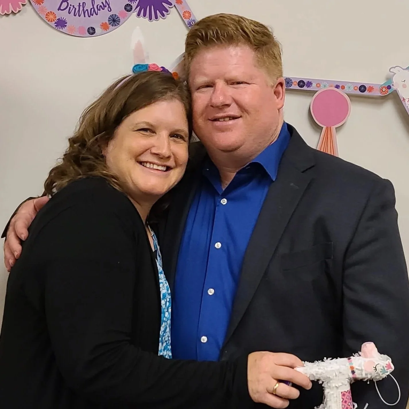 A smiling woman and man hugging at a birthday celebration with pink and purple decorations in the background.