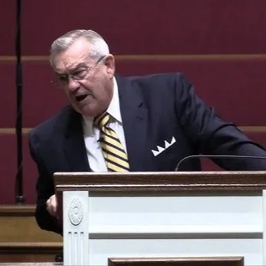 A man in a dark suit and striped tie speaking at a podium, appearing to be expressive during a speech or debate.