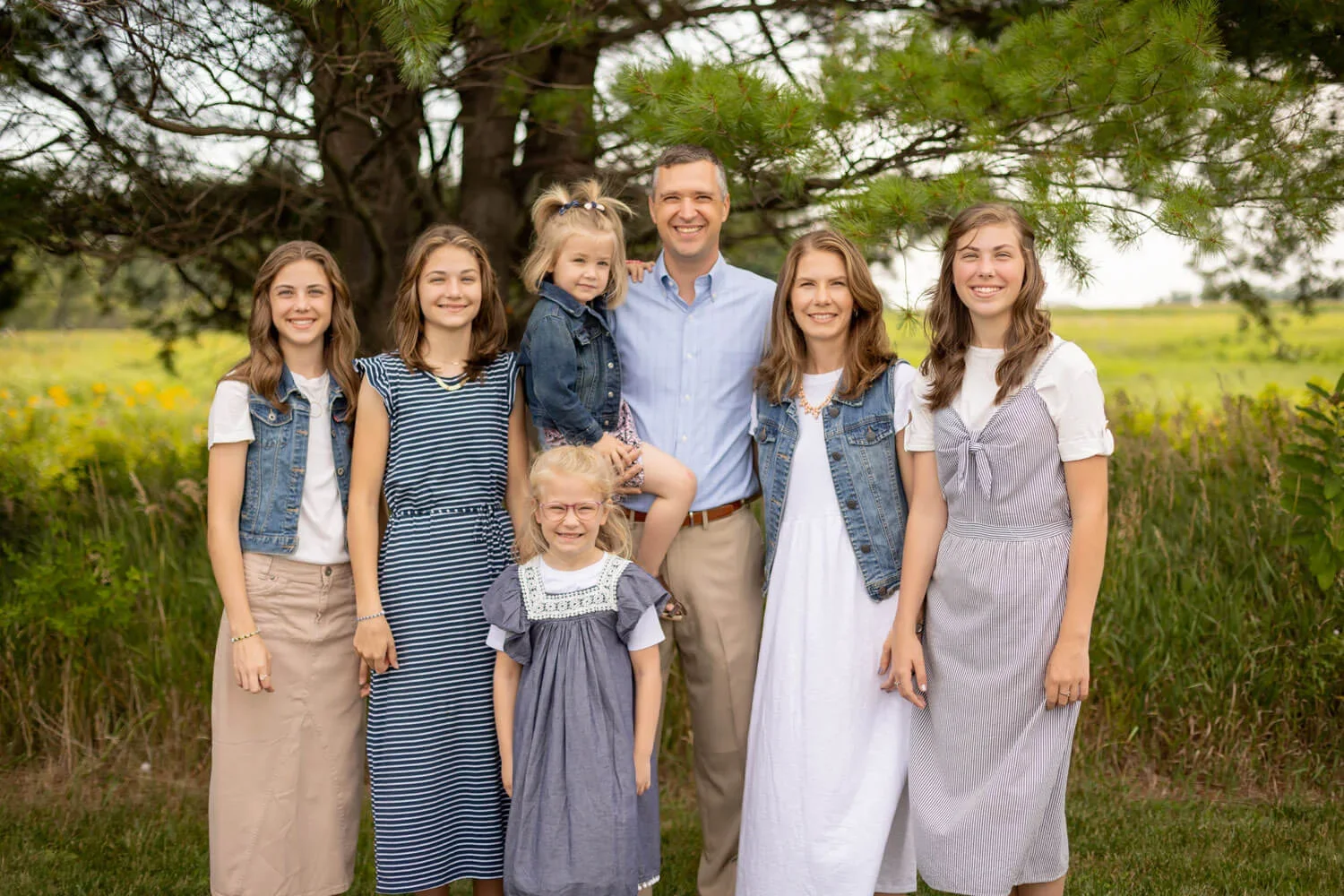 Family photo outdoors under a large tree, featuring six females and one male, all smiling at the camera.
