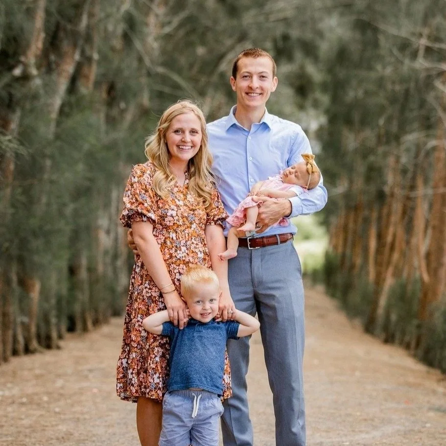 A family of four posing outdoors on a tree-lined dirt path. The father is holding a baby girl, and a young boy is standing in front of them. The mother stands beside the father, all smiling at the camera.