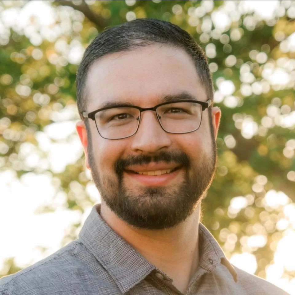 A smiling man with glasses and a beard, outdoors with blurred trees and sunlight in the background.