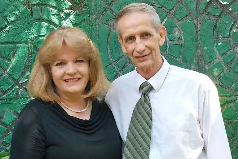 A woman with shoulder-length blonde hair wearing a black top and pearl necklace, standing next to an older man with short gray hair in a white shirt and patterned tie, in front of a green mosaic wall.