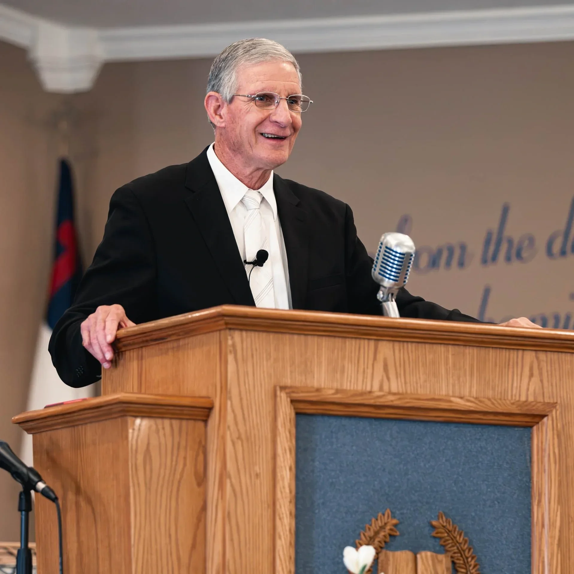 An elderly man with gray hair and glasses speaking at a wooden podium with a vintage microphone, wearing a black suit, white shirt, and white tie, in a church setting.