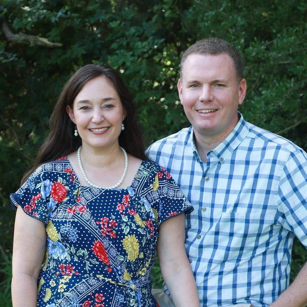 A woman and man standing outdoors in front of green foliage, smiling for the camera.