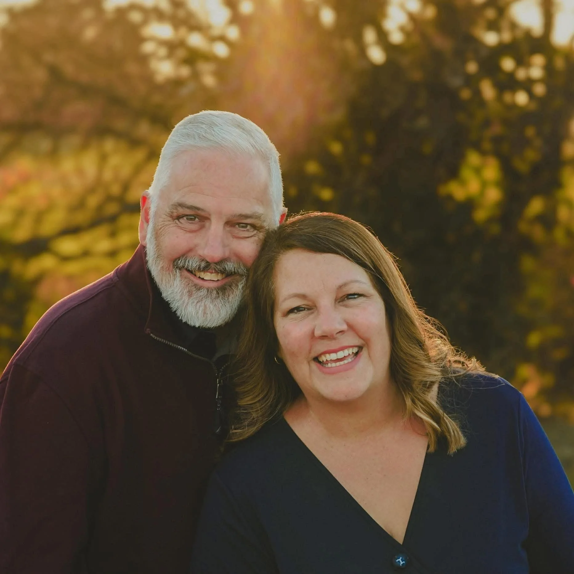 A happy middle-aged couple smiling outdoors during sunset with autumn trees in the background.