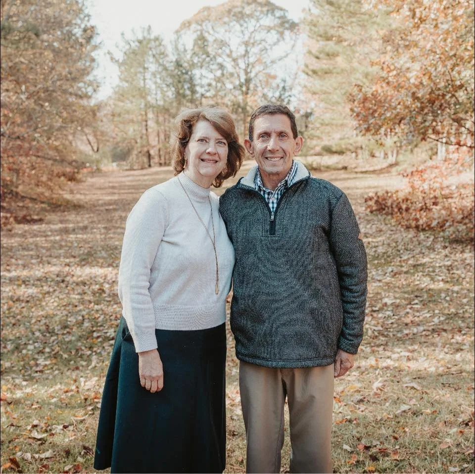 A woman and a man standing together outdoors in an autumn setting with trees and fallen leaves, smiling at the camera.