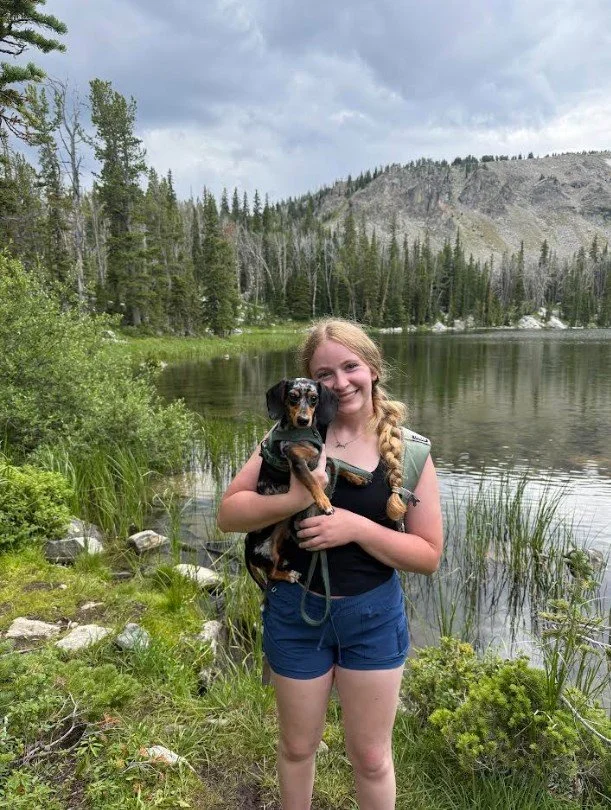Young woman holding a small black and tan dog by a lake surrounded by trees and mountains.