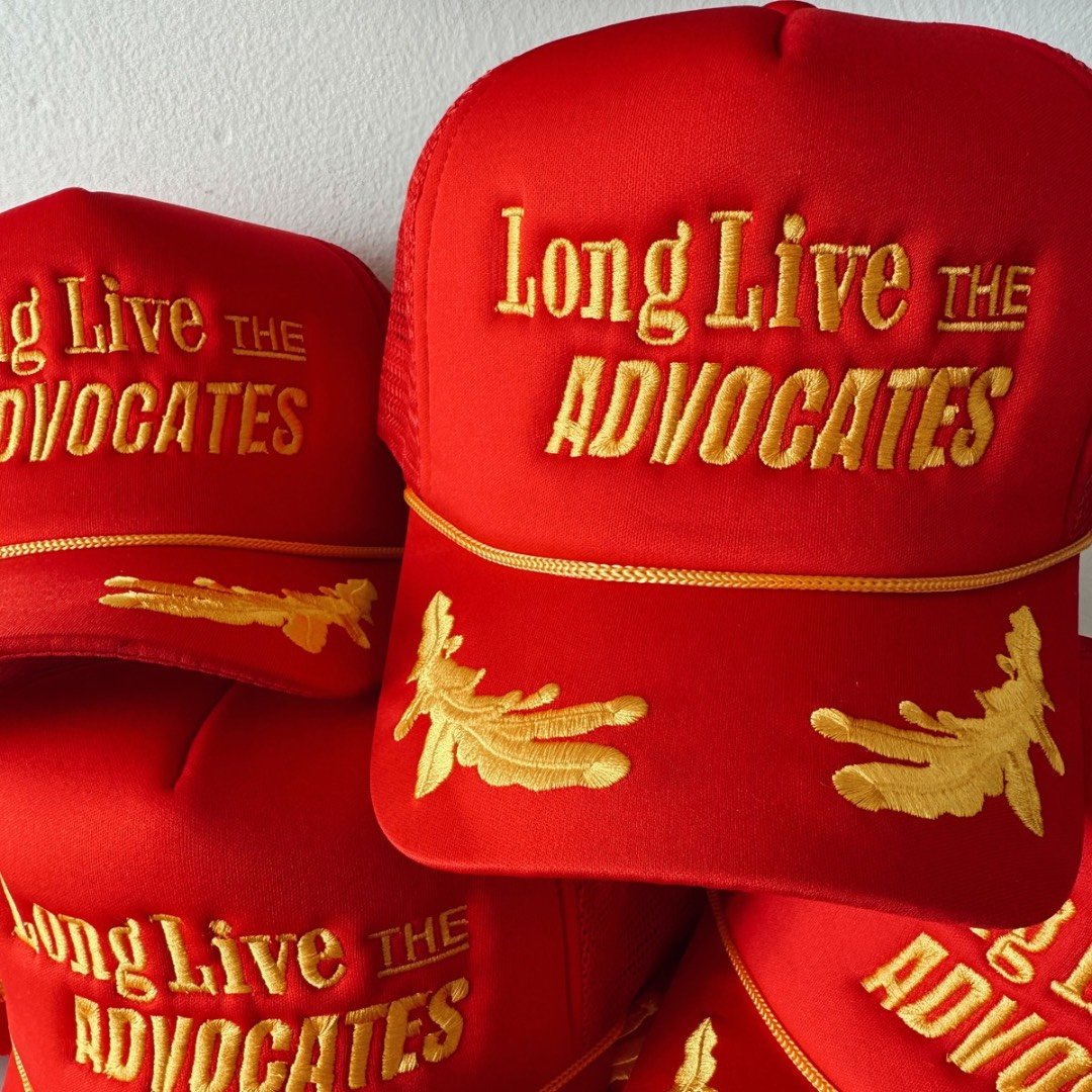 Red trucker hat with yellow embroidery that reads 'Long Live the Advocates' and decorative yellow oak leaf embroidery on the brim.