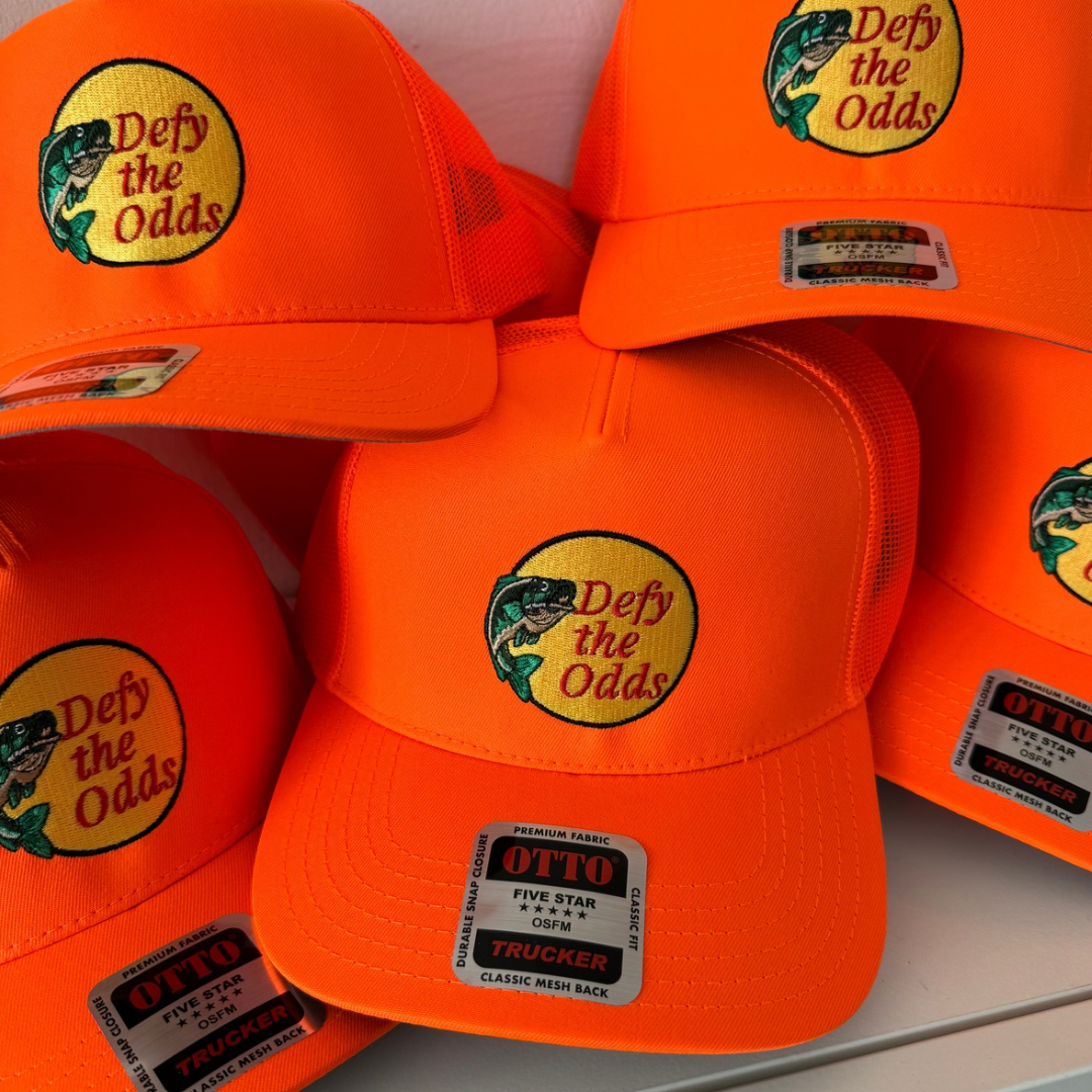 Otto bright orange trucker caps with a logo and slogan, displaying a fish graphic and the text "Defy the Odds."