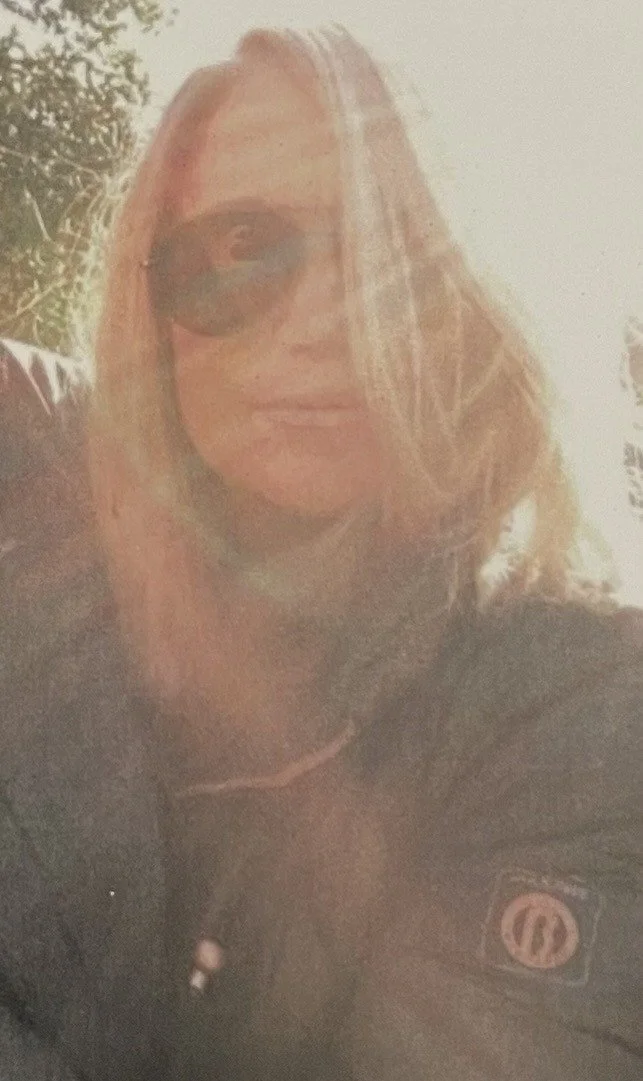 A woman with blonde hair wearing sunglasses and a dark jacket outdoors in sunlight.