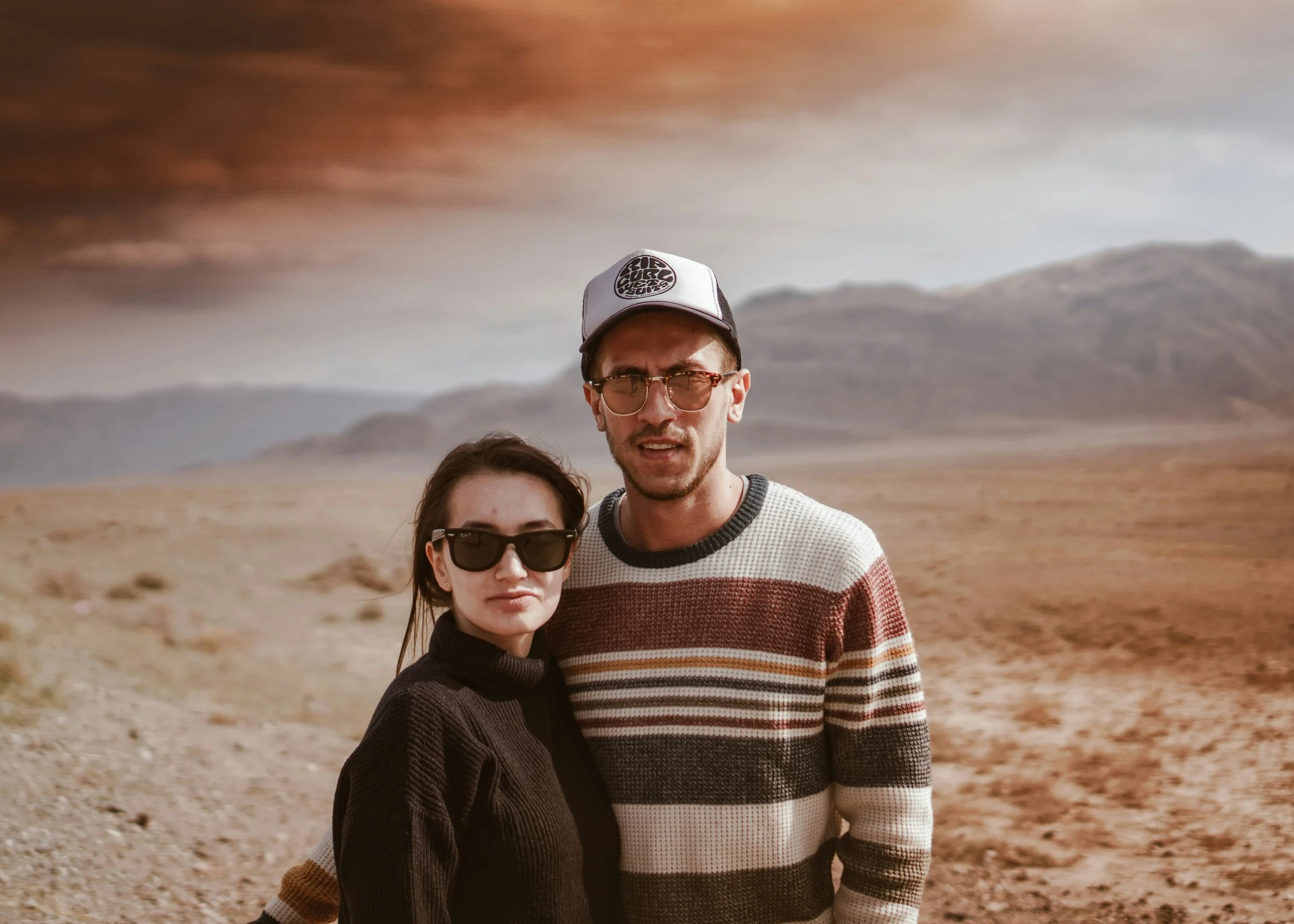 A man and woman standing close together outdoors in a desert landscape with mountains in the background, both wearing sunglasses and casual clothing.
