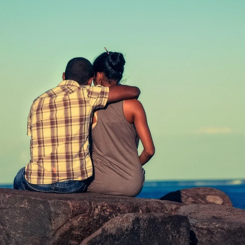 A boy and a girl sit on rocks by the water, with the boy hugging the girl from behind, under a clear sky.