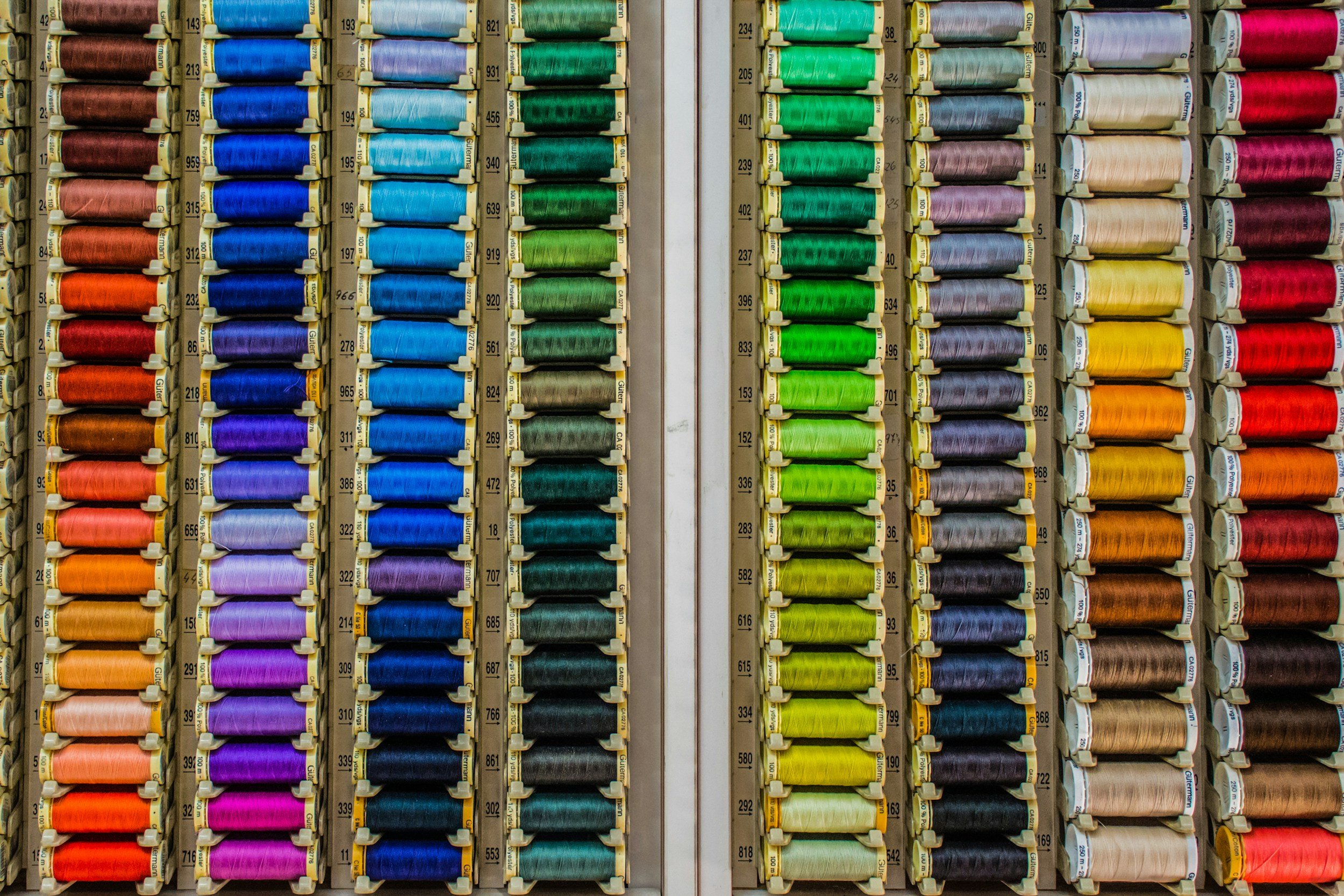 Close-up view of a wall of colorful thread spools organized in columns by color, including red, orange, pink, purple, blue, green, gray, yellow, and brown.