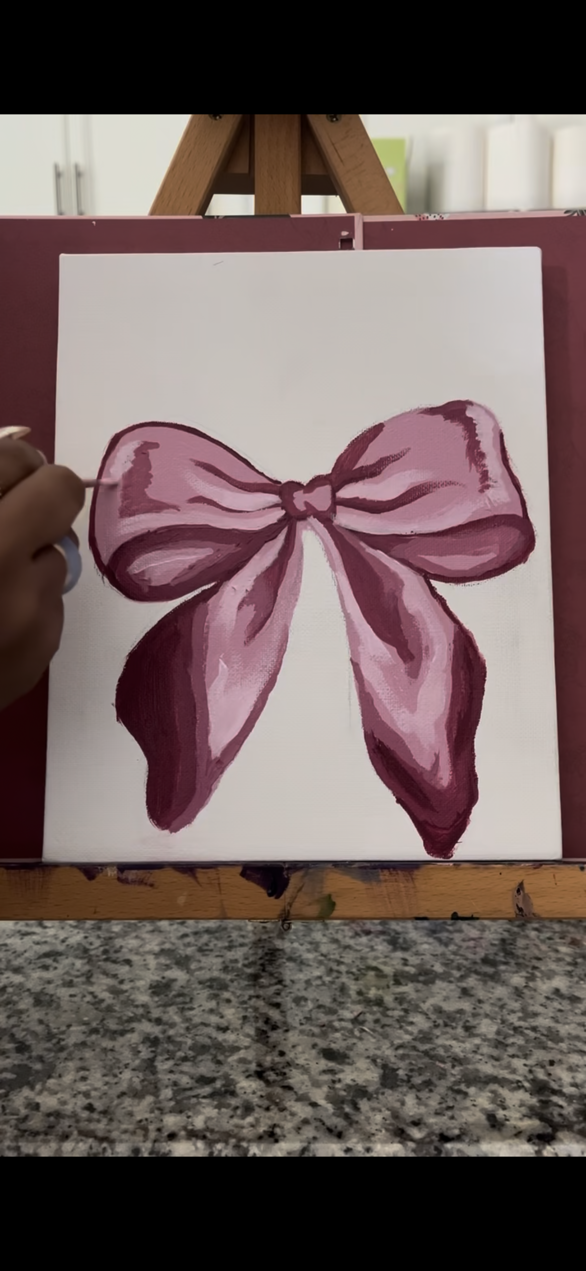 Person painting a pink bow on canvas with a light blue brush.