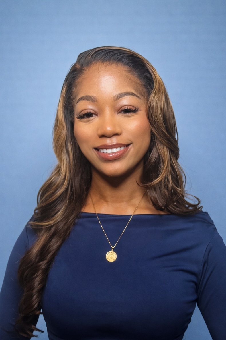 Professional headshot of a young woman with long wavy brown hair and blonde highlights wearing a navy blue top and gold pendant necklace against a clean blue background.