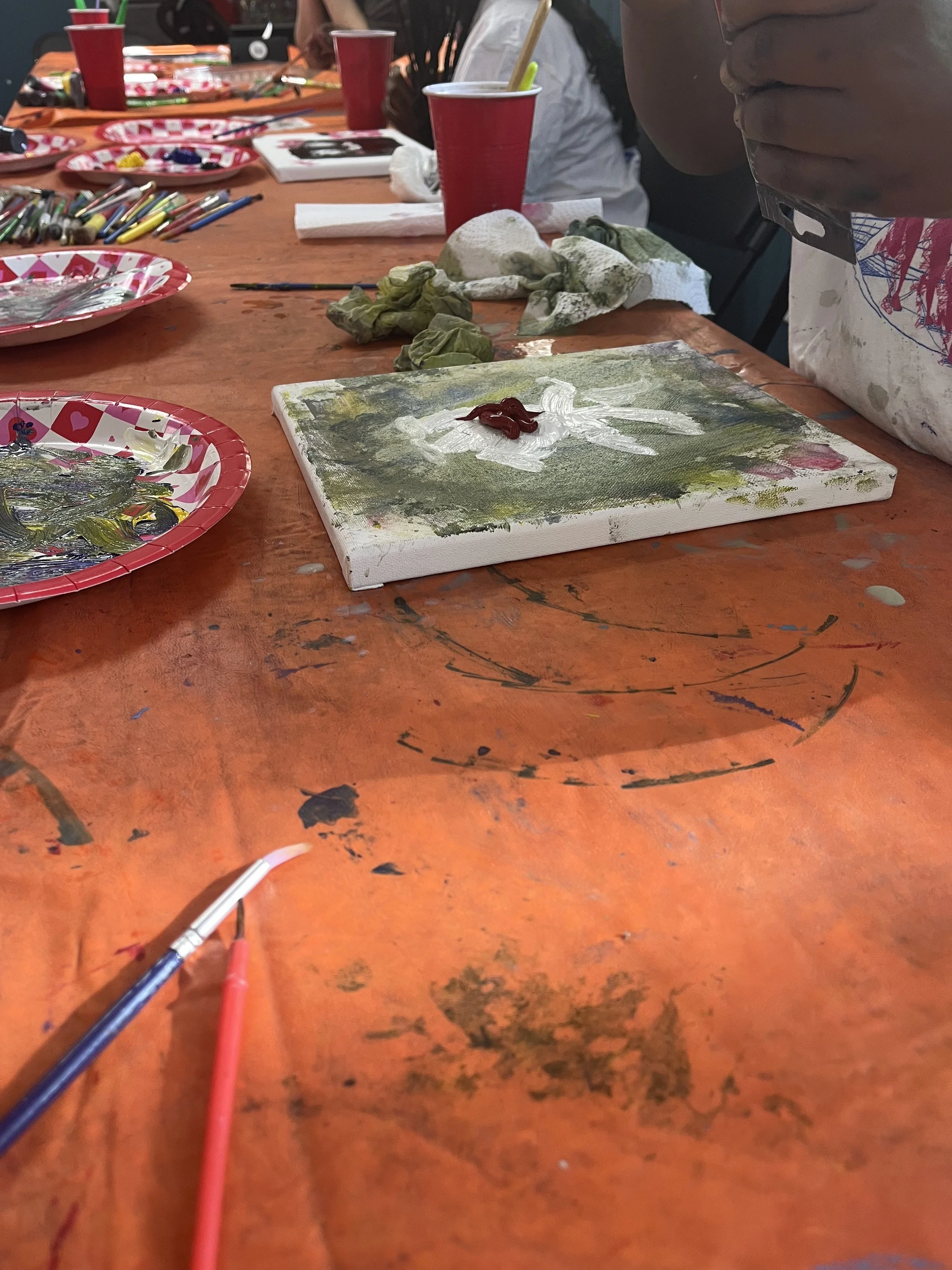 Close-up of an arts and crafts workspace with a small canvas featuring a painted flower, surrounded by paint smudges and used tissues. There are red cups, paintbrushes, and paint palettes with leftover paint on the disorganized, paint-stained table.